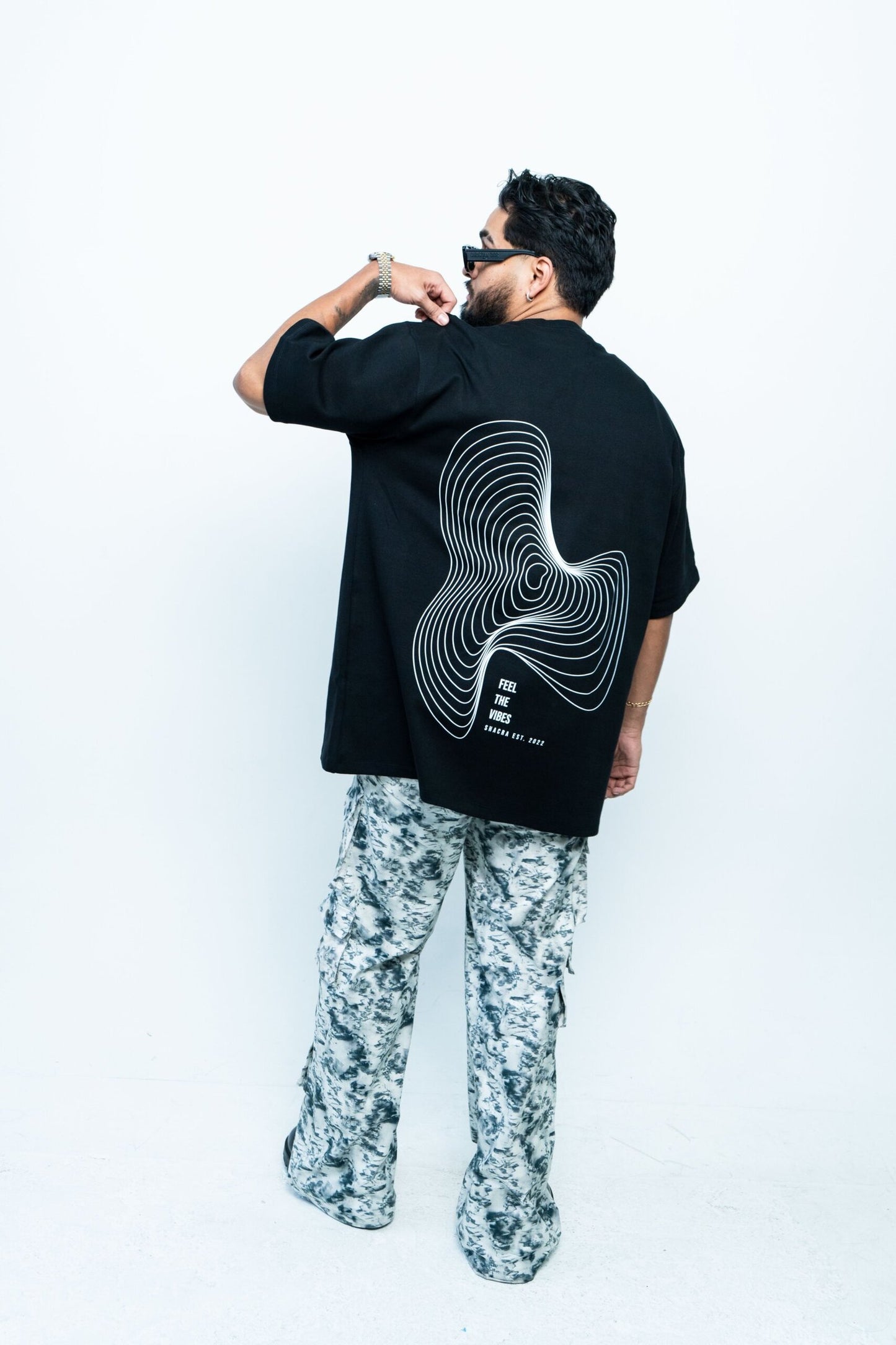 Modern man wearing a black oversized graphic t-shirt with a swirling line art design and text, paired with patterned cargo pants, stylish accessories, and sunglasses, showcasing casual streetwear fashion.