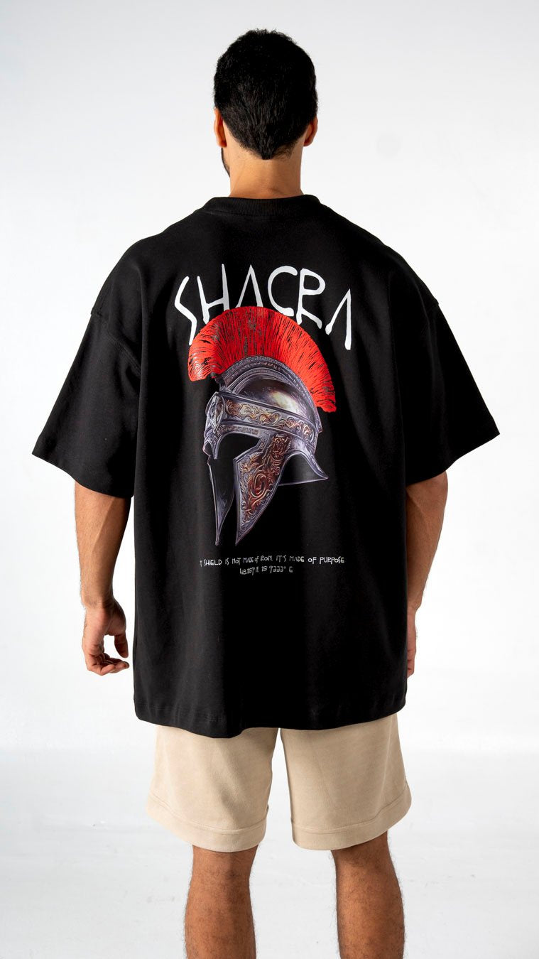 Black graphic t-shirt featuring a detailed Spartan helmet with red crest, bold "SHACRA" print, and motivational quote, styled for casual wear, emphasizing streetwear fashion and urban apparel.