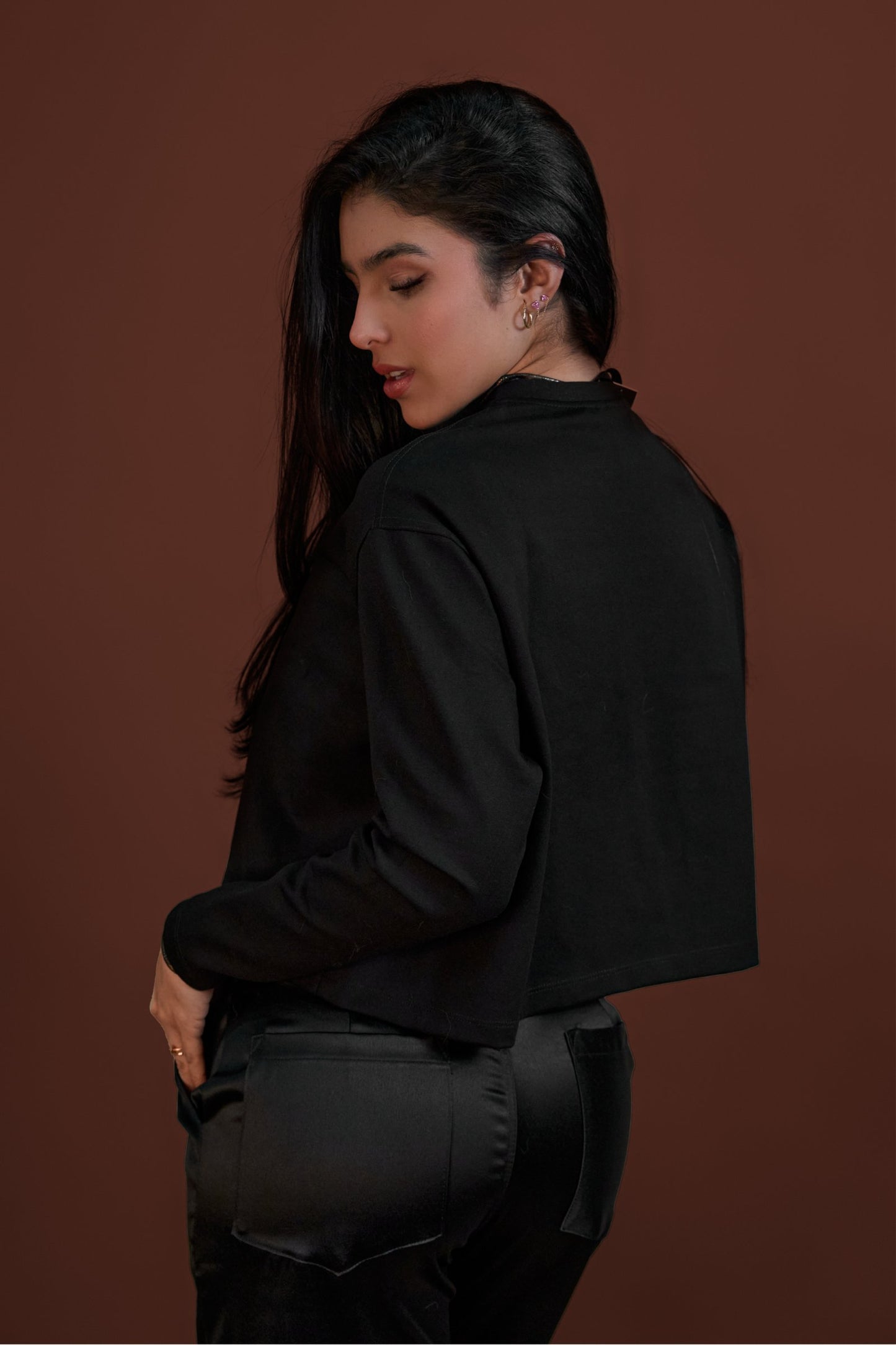 Black cropped jacket and matching leather pants worn by a woman with dark hair and earrings, posing against a warm brown background.
