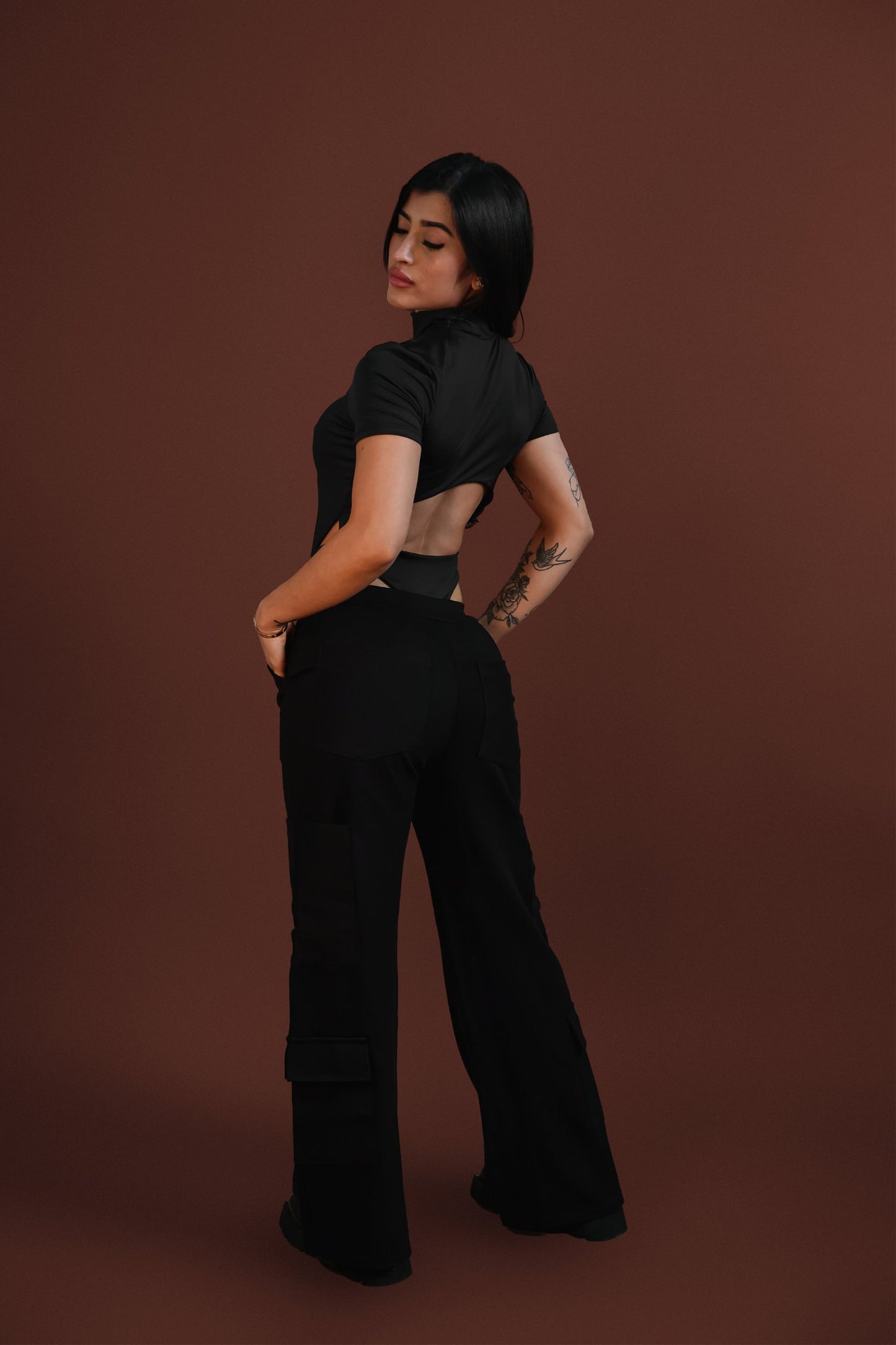 Elegant woman in black crop top and cargo pants, showcasing stylish streetwear fashion with tattoos, against a warm brown background. Perfect for urban fashion, accessories, and modern style inspiration.