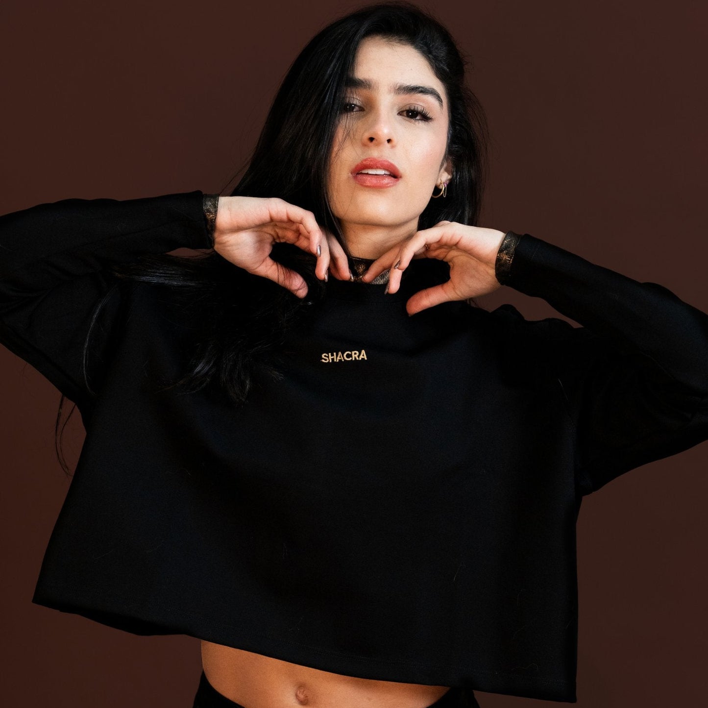 Vibrant woman modeling in stylish black casual wear, posing confidently with hands near her face against a solid brown background, showcasing fashion, confidence, and modern style.