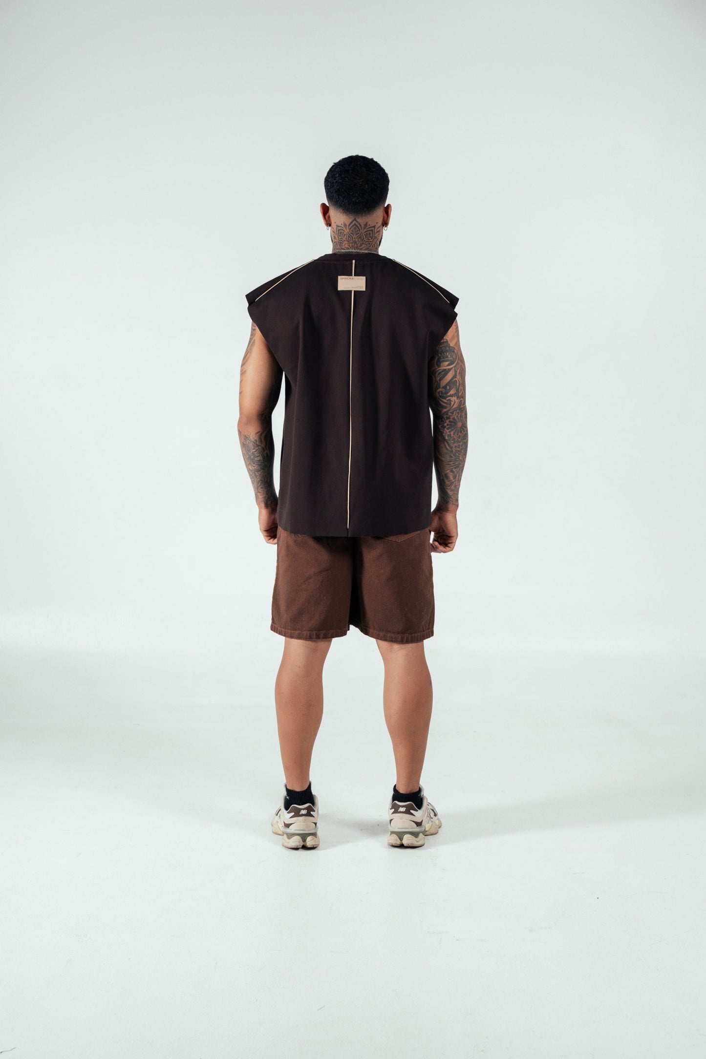 Modern fashion model showcasing contemporary streetwear with a sleeveless black top, brown shorts, and sneakers in a minimal studio setting.