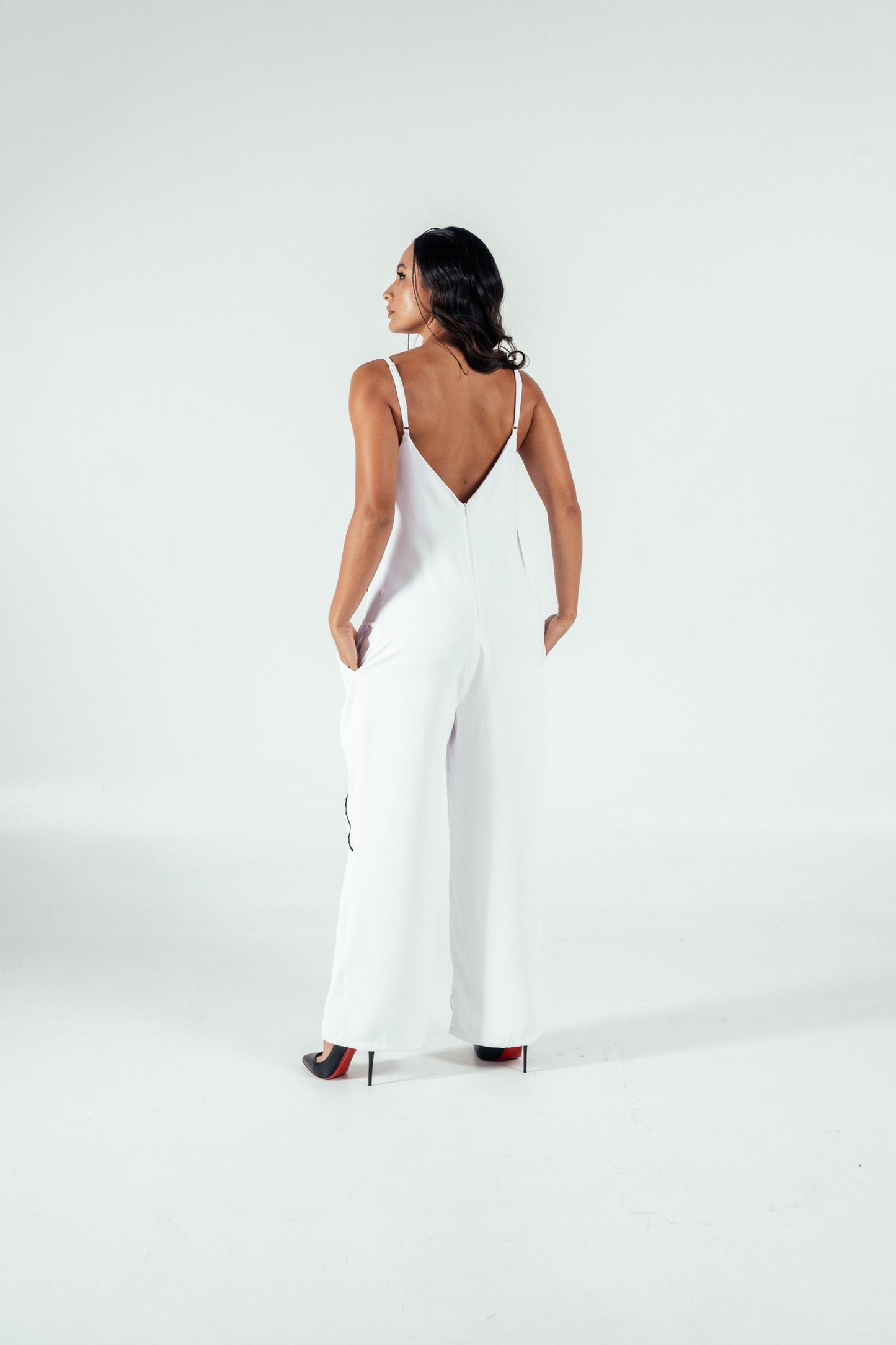 Elegant woman in a white stylish jumpsuit with a deep V back, standing on high heels against a neutral background, showcasing modern fashion and sophisticated style.