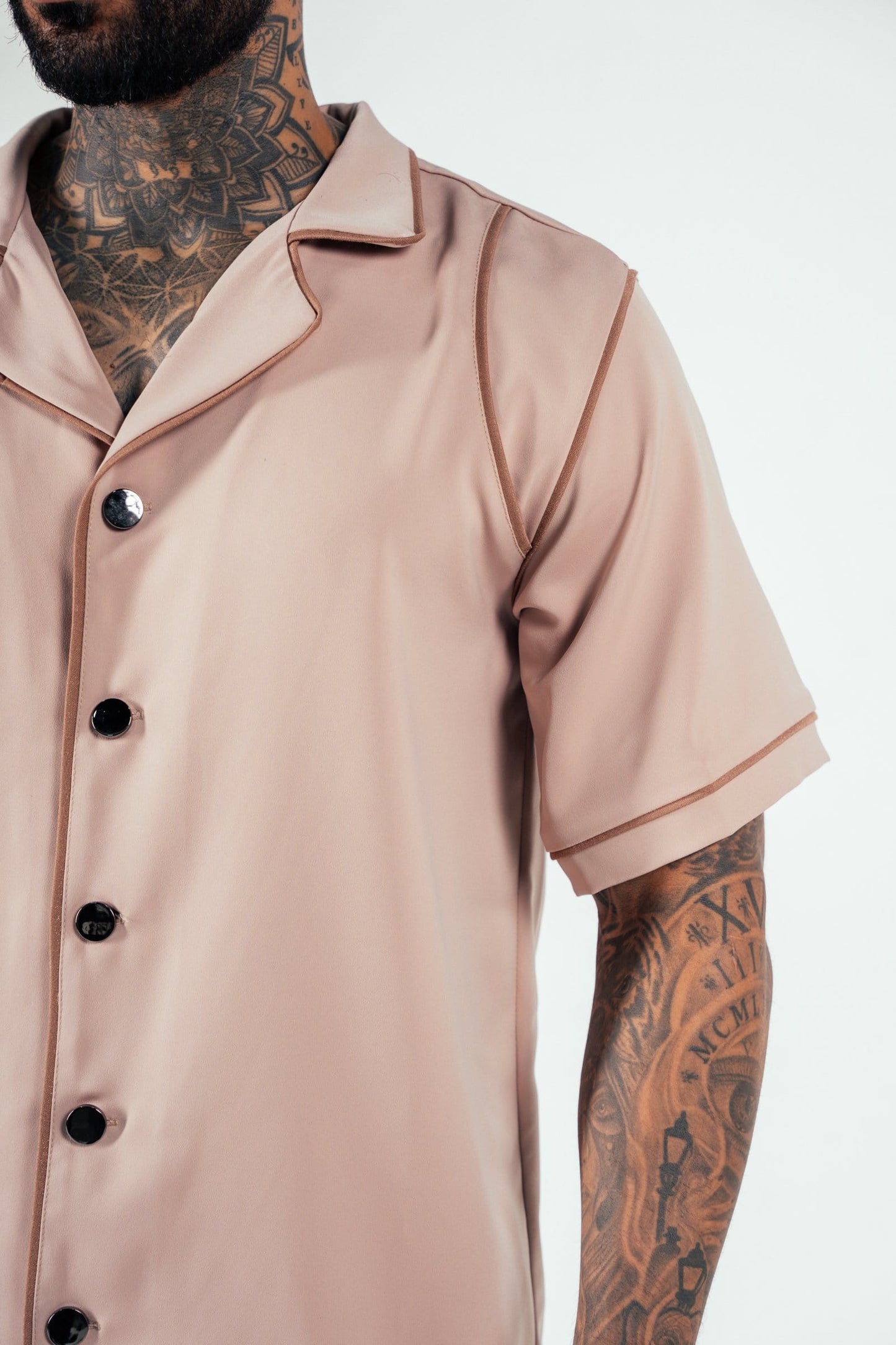 Bold tattooed man wearing a beige satin short-sleeve button-up shirt, showcasing elegant fashion and tattoo artistry, with a minimalist background emphasizing style and details.