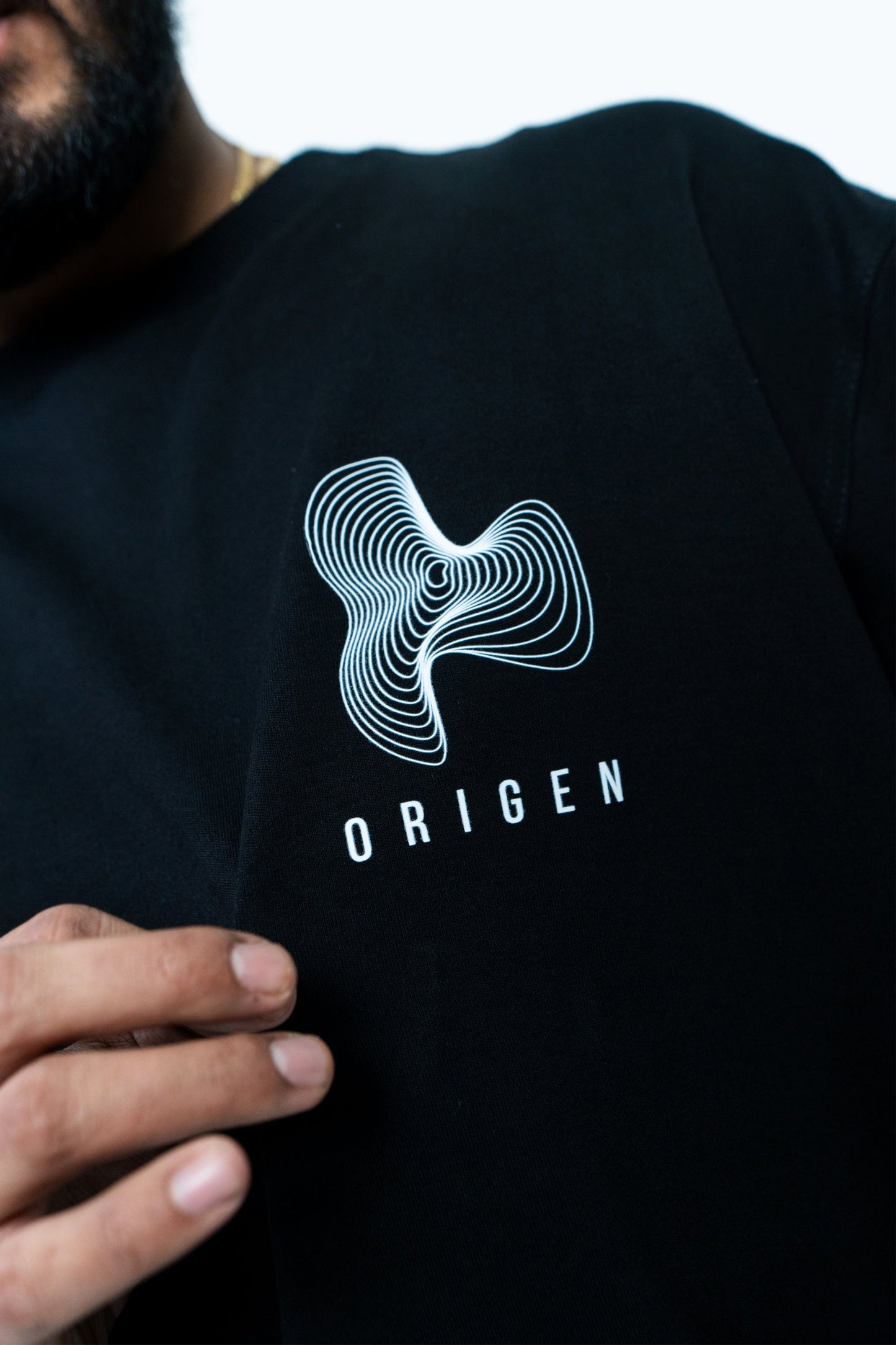 Aesthetic black t-shirt with white ORIGEN logo featuring a unique line art design, worn by a person with a partial view of their face and hand, emphasizing modern style and brand identity.