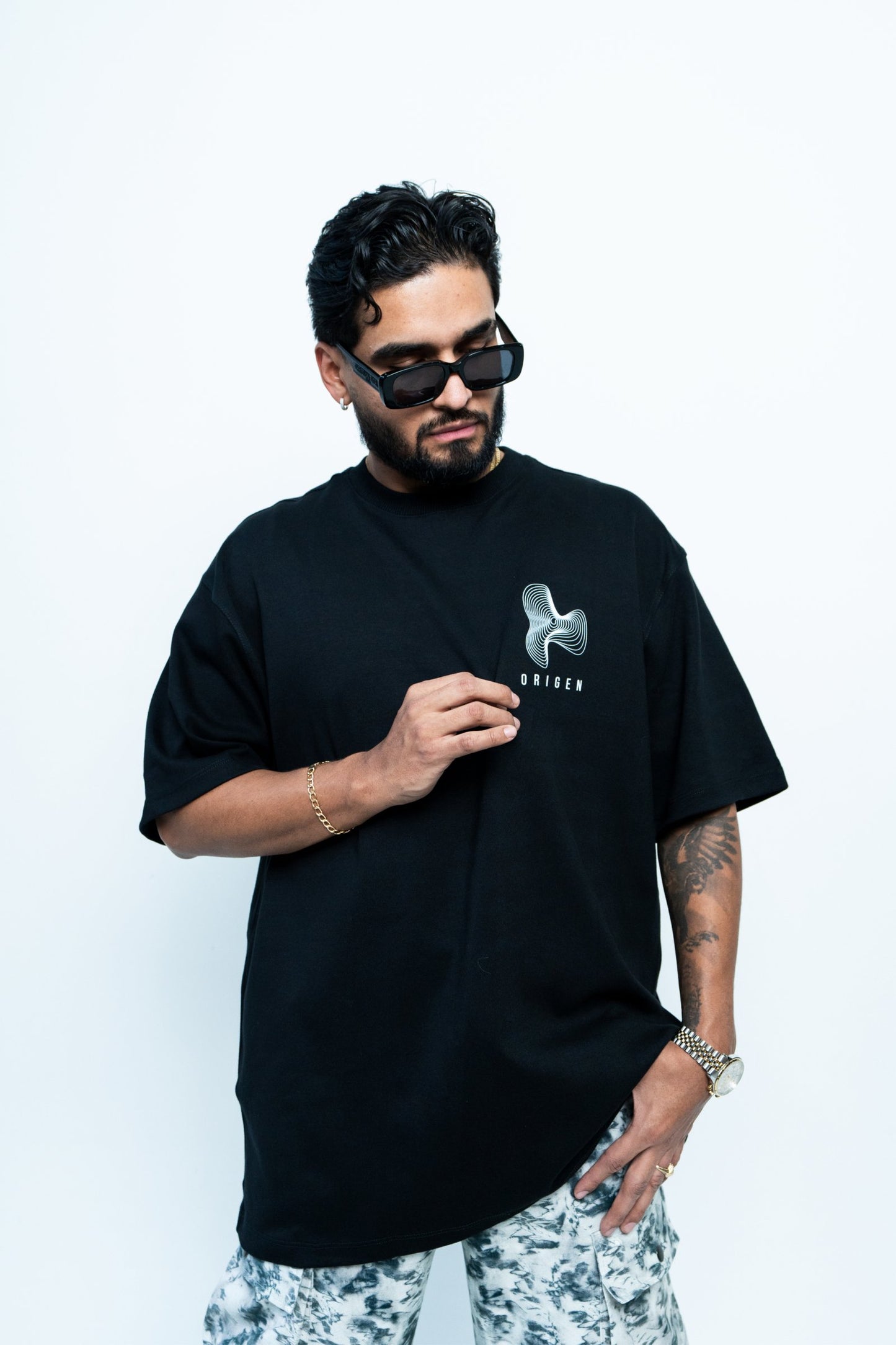 Modern male fashion model wearing black graphic t-shirt, sunglasses, and patterned pants, showcasing trendy streetwear style and accessories for fashion-forward urban look.