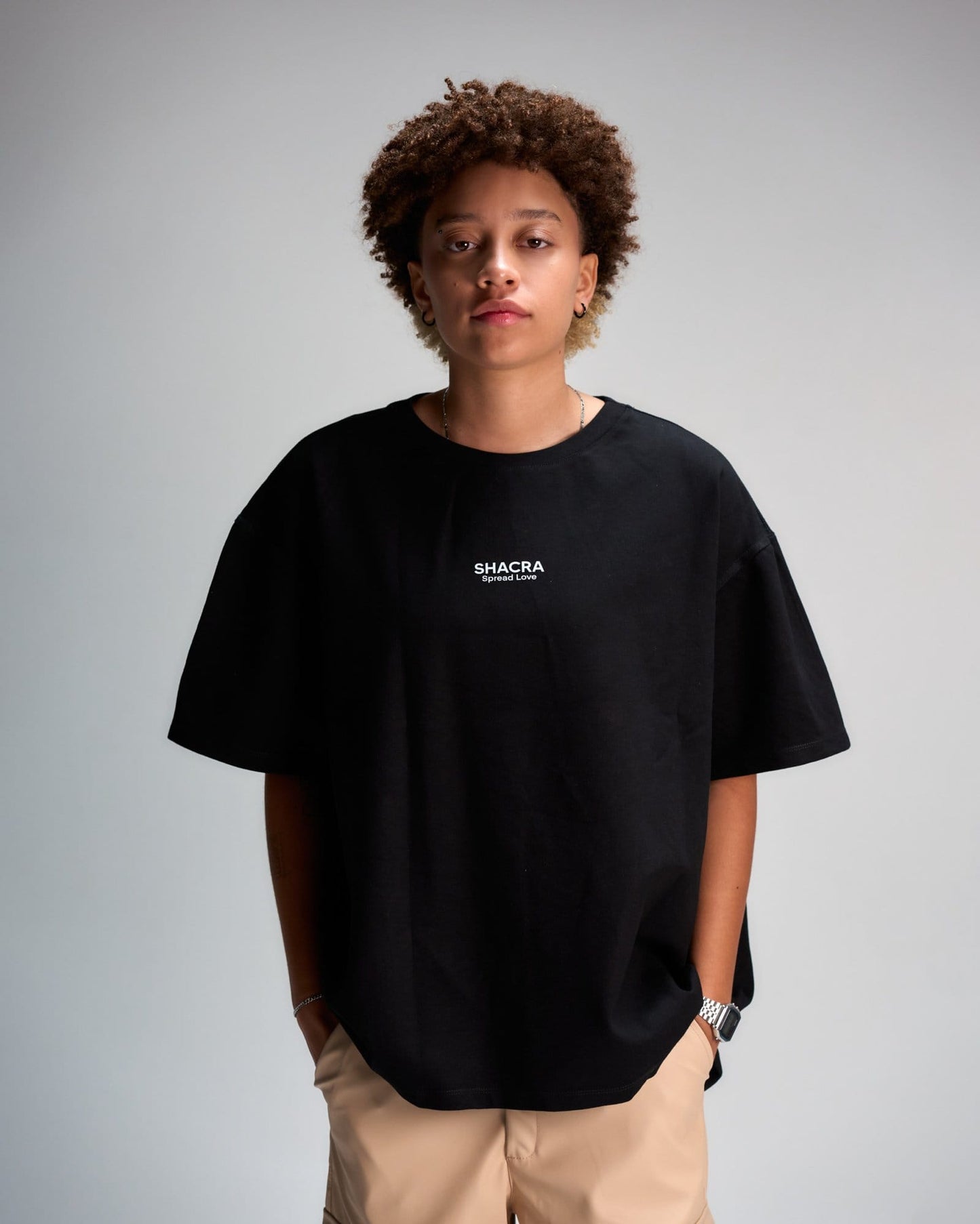Black oversized T-shirt with "SHACRA Spread Love" logo, worn by a young woman with natural curly hair, standing against a plain grey background. Casual fashion styling with beige pants, accessorized with jewelry and a watch.