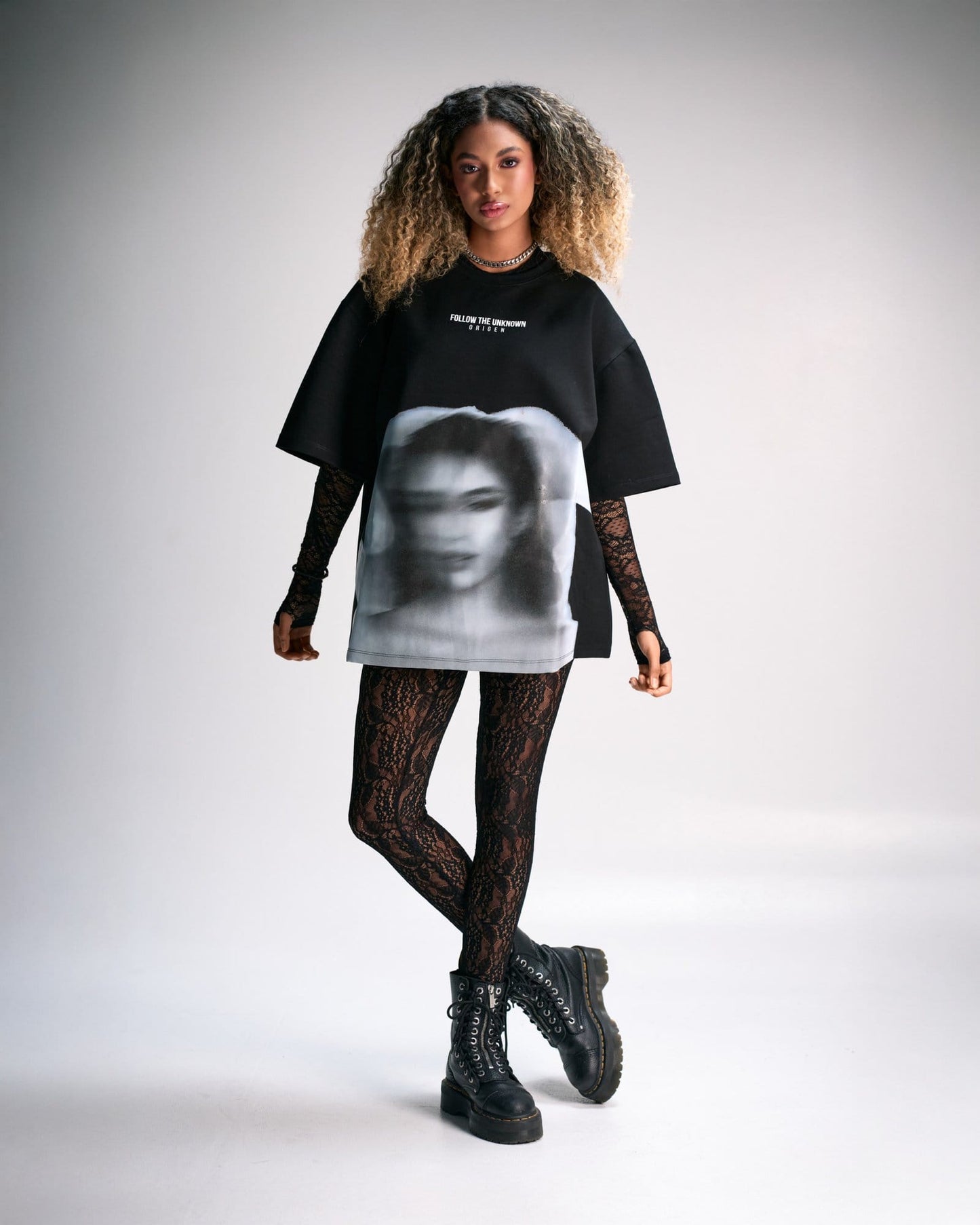 Bold fashion-forward young woman wearing an oversized black graphic t-shirt, lace gloves, patterned leggings, and chunky combat boots, showcasing edgy street style and modern urban fashion.