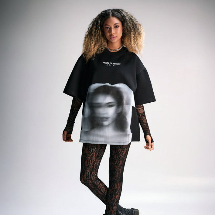 Bold fashion-forward young woman wearing an oversized black graphic t-shirt, lace gloves, patterned leggings, and chunky combat boots, showcasing edgy street style and modern urban fashion.