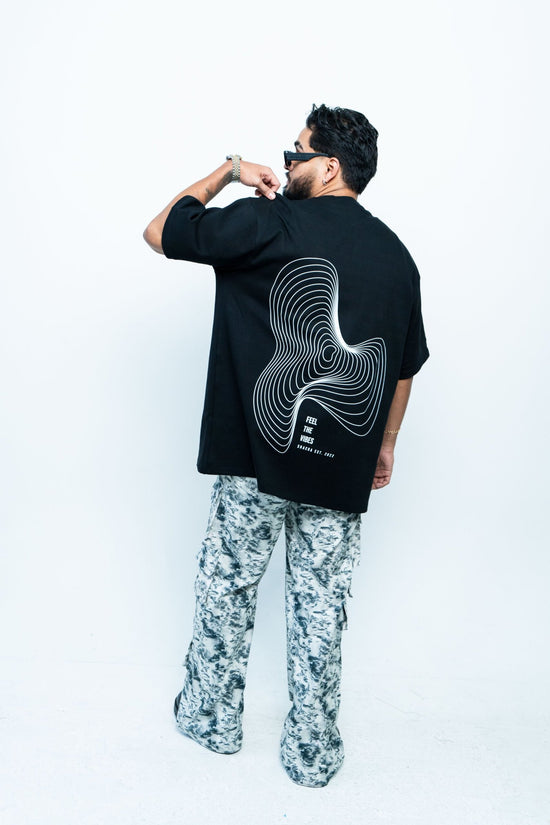 Modern man wearing a black oversized graphic t-shirt with a swirling line art design and text, paired with patterned cargo pants, stylish accessories, and sunglasses, showcasing casual streetwear fashion.