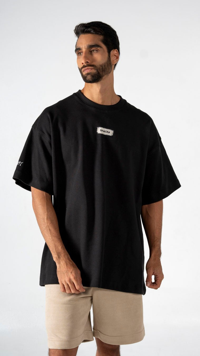 A black oversized T-shirt with a small Shacra logo patch on the chest, featuring a relaxed fit and casual style, paired with beige shorts, showcasing modern streetwear fashion.