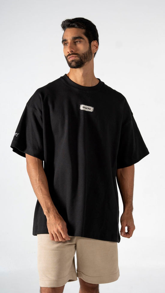 A black oversized T-shirt with a small Shacra logo patch on the chest, featuring a relaxed fit and casual style, paired with beige shorts, showcasing modern streetwear fashion.