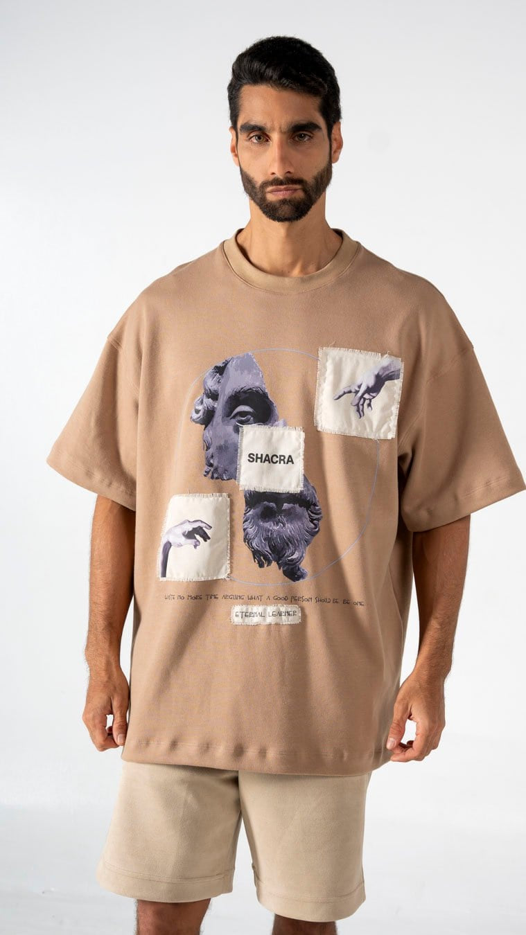 A man wearing a beige graphic T-shirt with artistic face and hand illustrations, featuring the brand name Shacra, against a plain gray background.