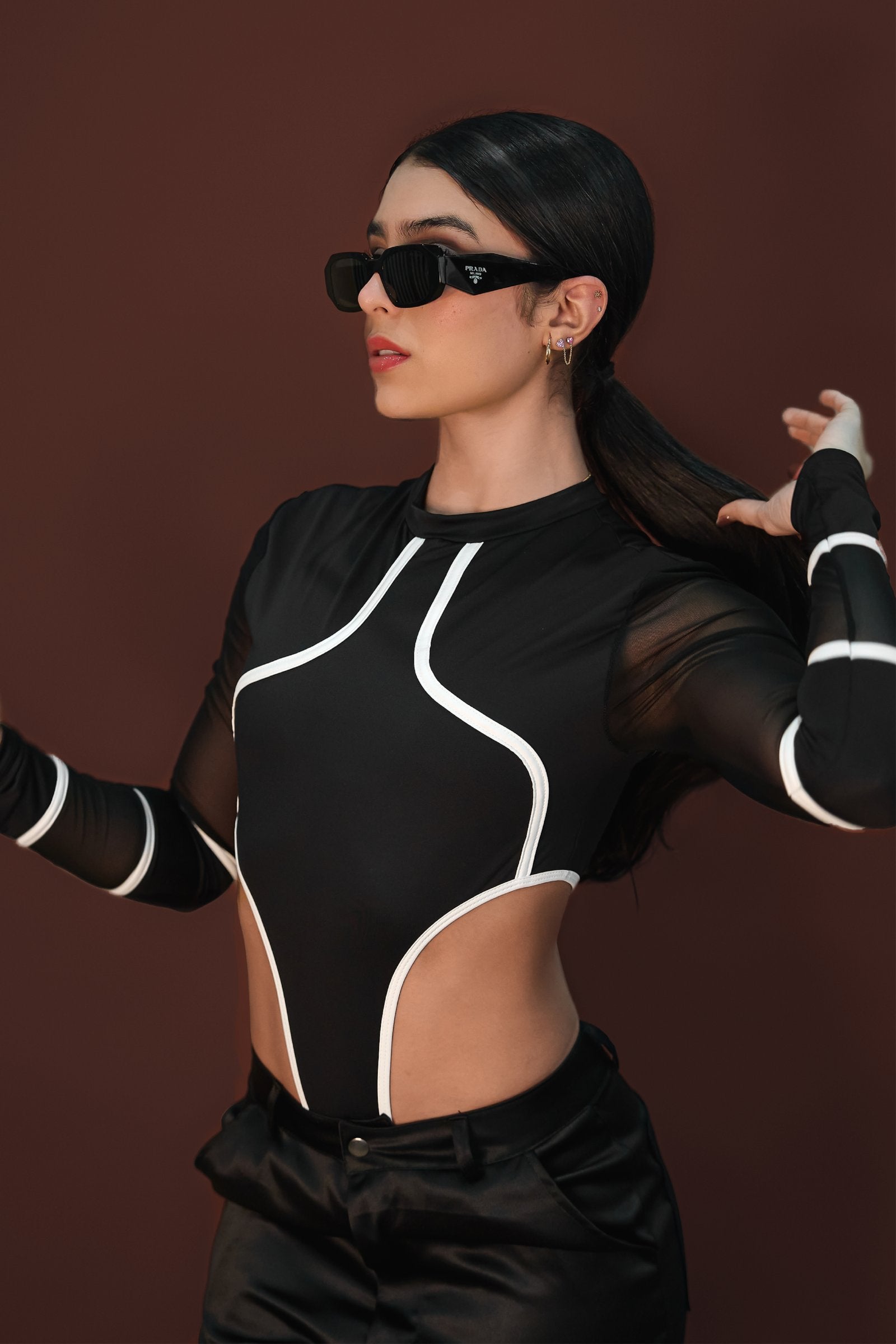 Bold fashion model wearing black Prada sunglasses, sporty black and white cut-out top, and black satin pants, posing confidently against a warm brown background.