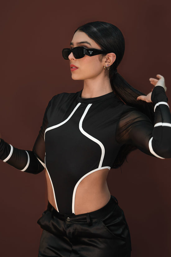 Bold fashion model wearing black Prada sunglasses, sporty black and white cut-out top, and black satin pants, posing confidently against a warm brown background.