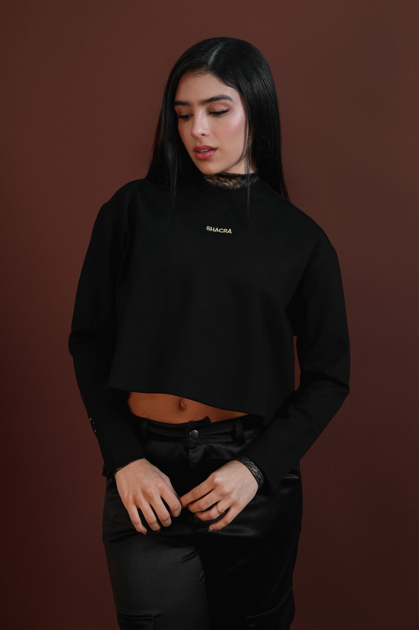 Stylish young woman wearing black sweatshirt with "Shacra" logo, posing against brown background for fashion and streetwear style photoshoot.