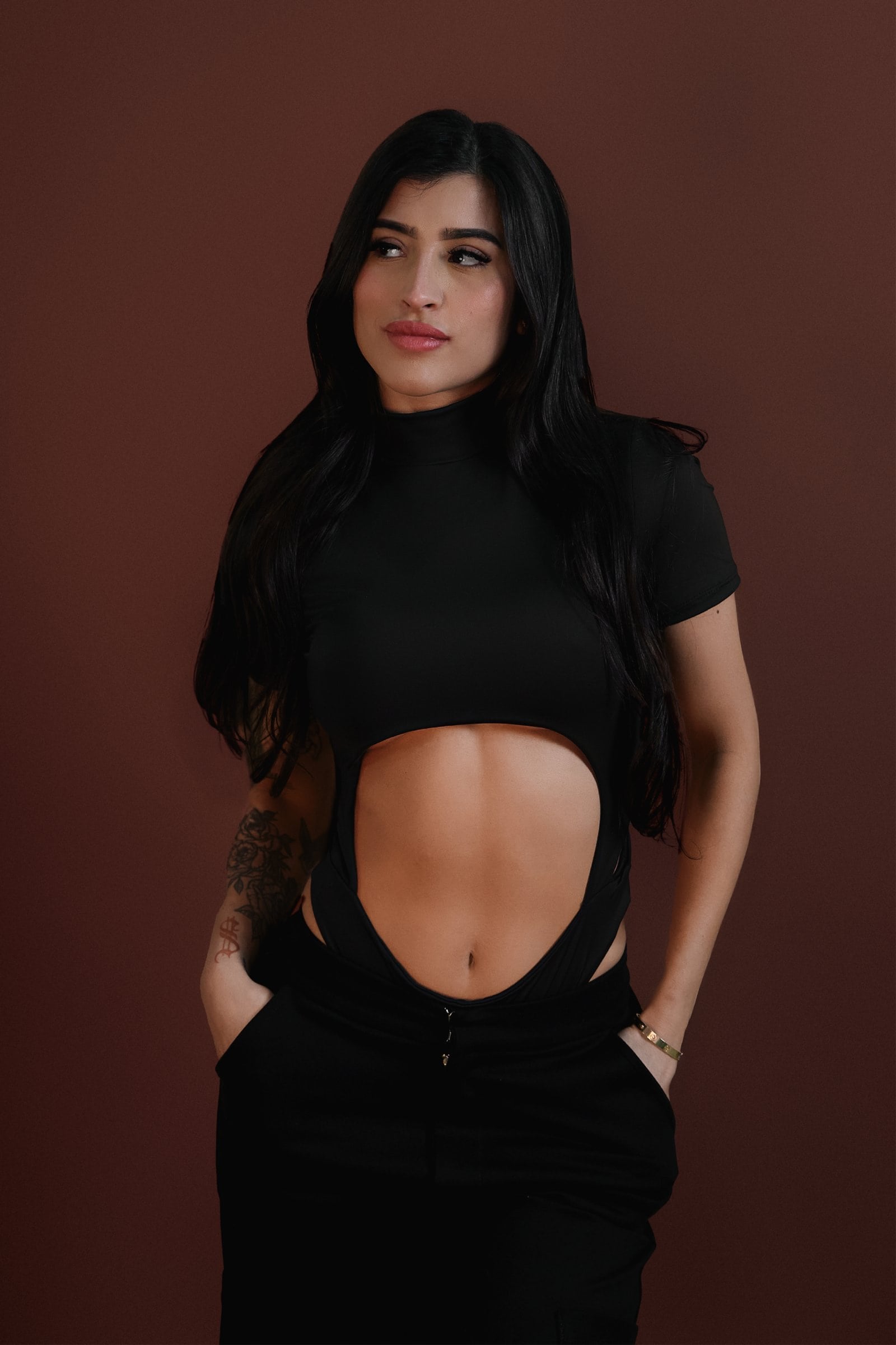Bold woman wearing a black cut-out top and black pants posing against a brown background, showcasing trendy fashion and confident style.