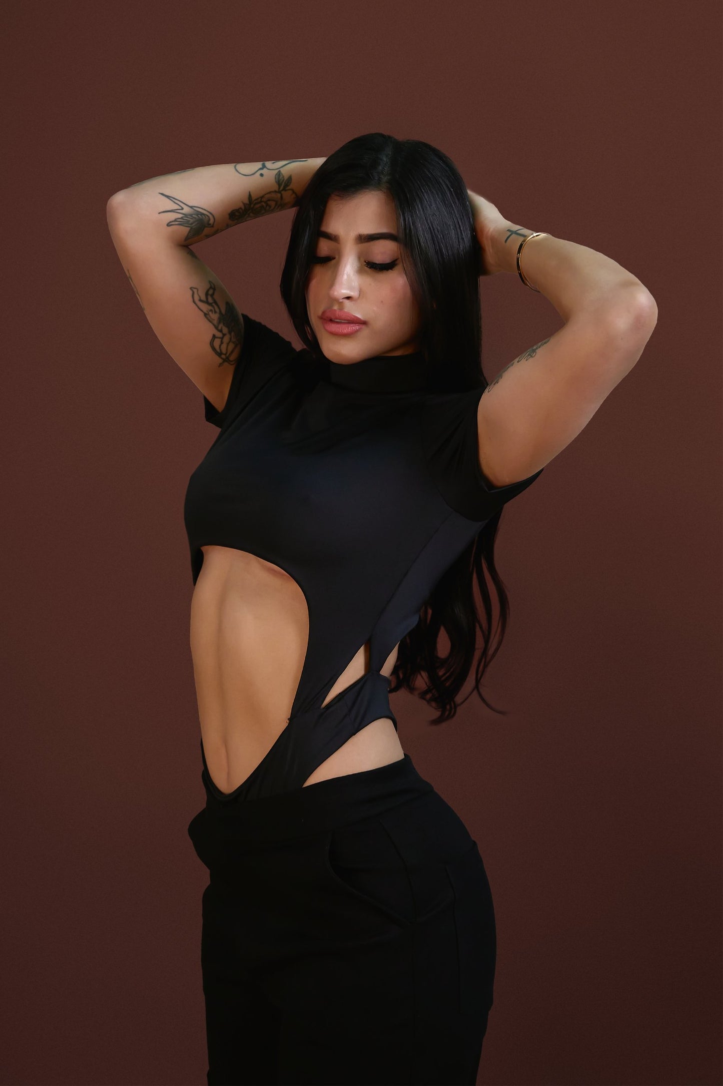 Black crop top with cutout design showing midriff, with tattoos on arms, dark hair, posing confidently against brown background.