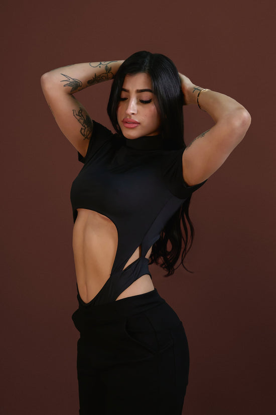 Black crop top with cutout design showing midriff, with tattoos on arms, dark hair, posing confidently against brown background.
