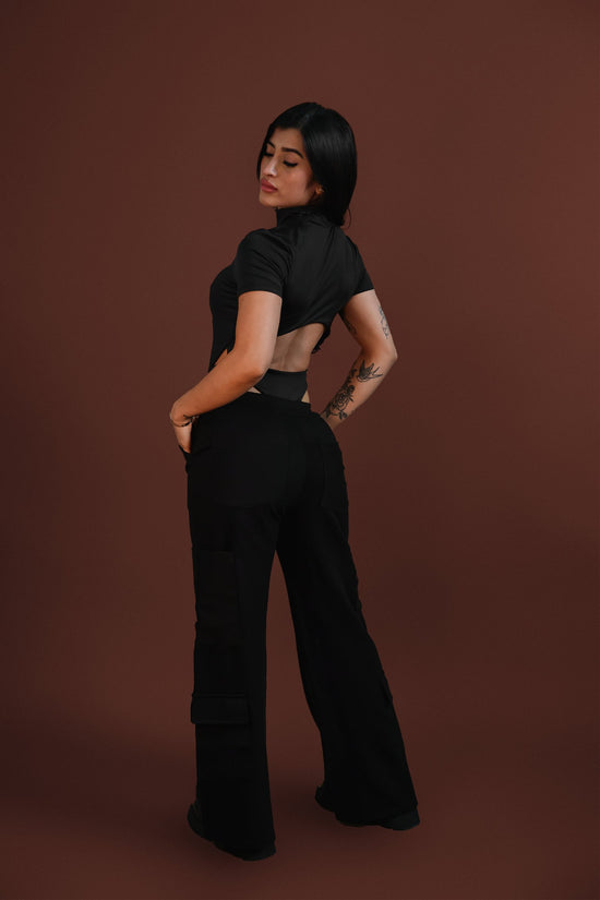 Elegant woman in black crop top and cargo pants, showcasing stylish streetwear fashion with tattoos, against a warm brown background. Perfect for urban fashion, accessories, and modern style inspiration.
