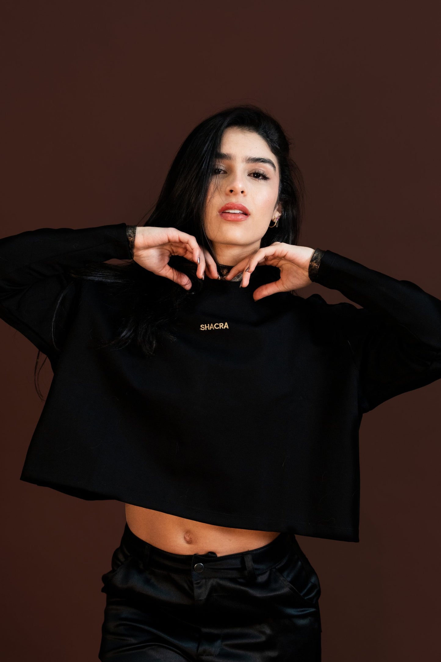 Vibrant woman modeling in stylish black casual wear, posing confidently with hands near her face against a solid brown background, showcasing fashion, confidence, and modern style.