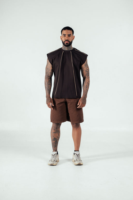 Bold tattooed man wearing a black sleeveless top with gold accents, brown shorts, and athletic sneakers, standing confidently in a minimal studio setting, showcasing modern streetwear fashion.
