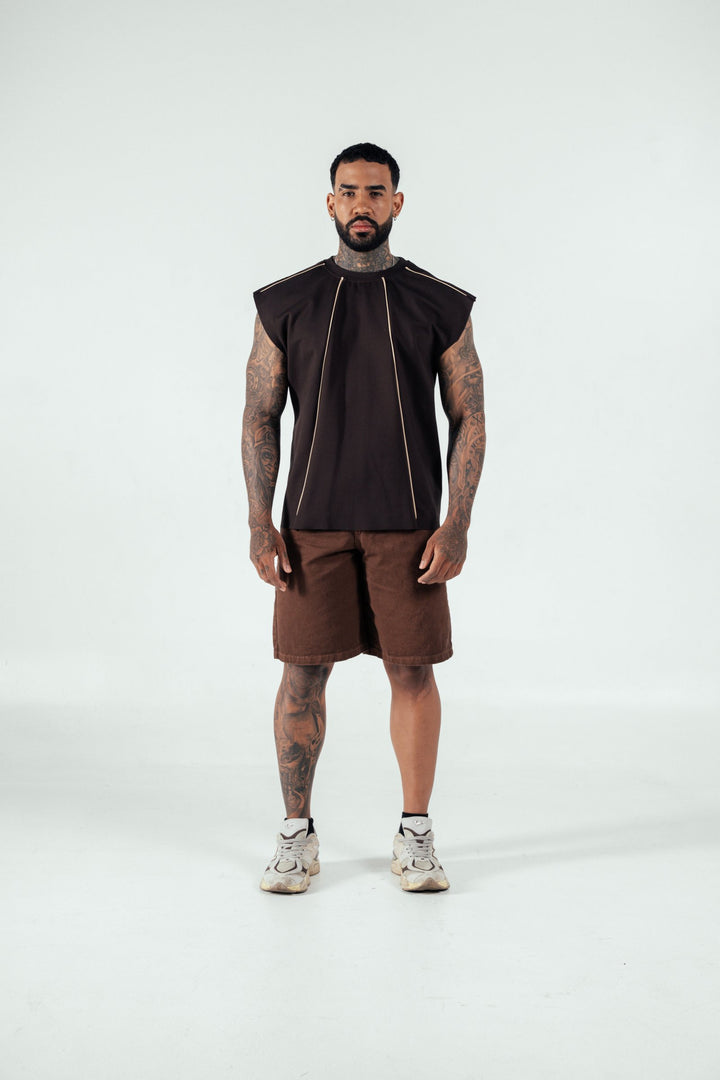 Bold tattooed man wearing a black sleeveless top with gold accents, brown shorts, and athletic sneakers, standing confidently in a minimal studio setting, showcasing modern streetwear fashion.