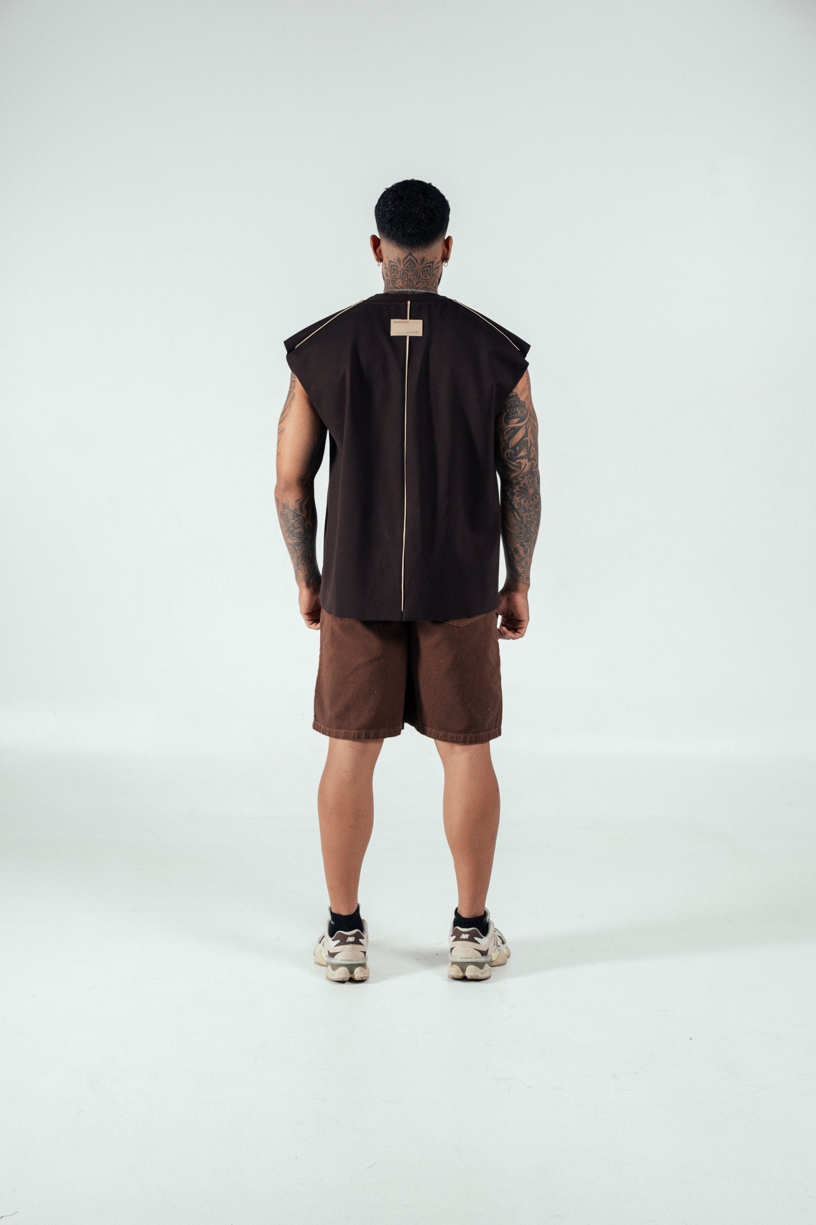 Modern fashion model showcasing contemporary streetwear with a sleeveless black top, brown shorts, and sneakers in a minimal studio setting.