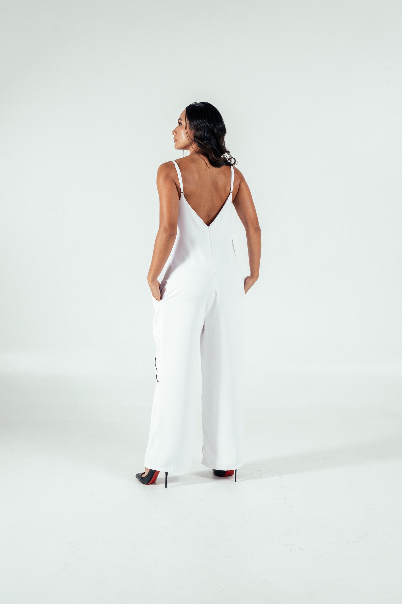 Elegant woman in a white stylish jumpsuit with a deep V back, standing on high heels against a neutral background, showcasing modern fashion and sophisticated style.