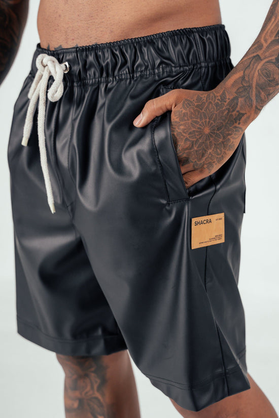 Black faux leather shorts with a drawstring waist and a small leather patch label, worn by a model with tattoos on their hand and leg. Perfect for stylish streetwear and casual summer outfits.