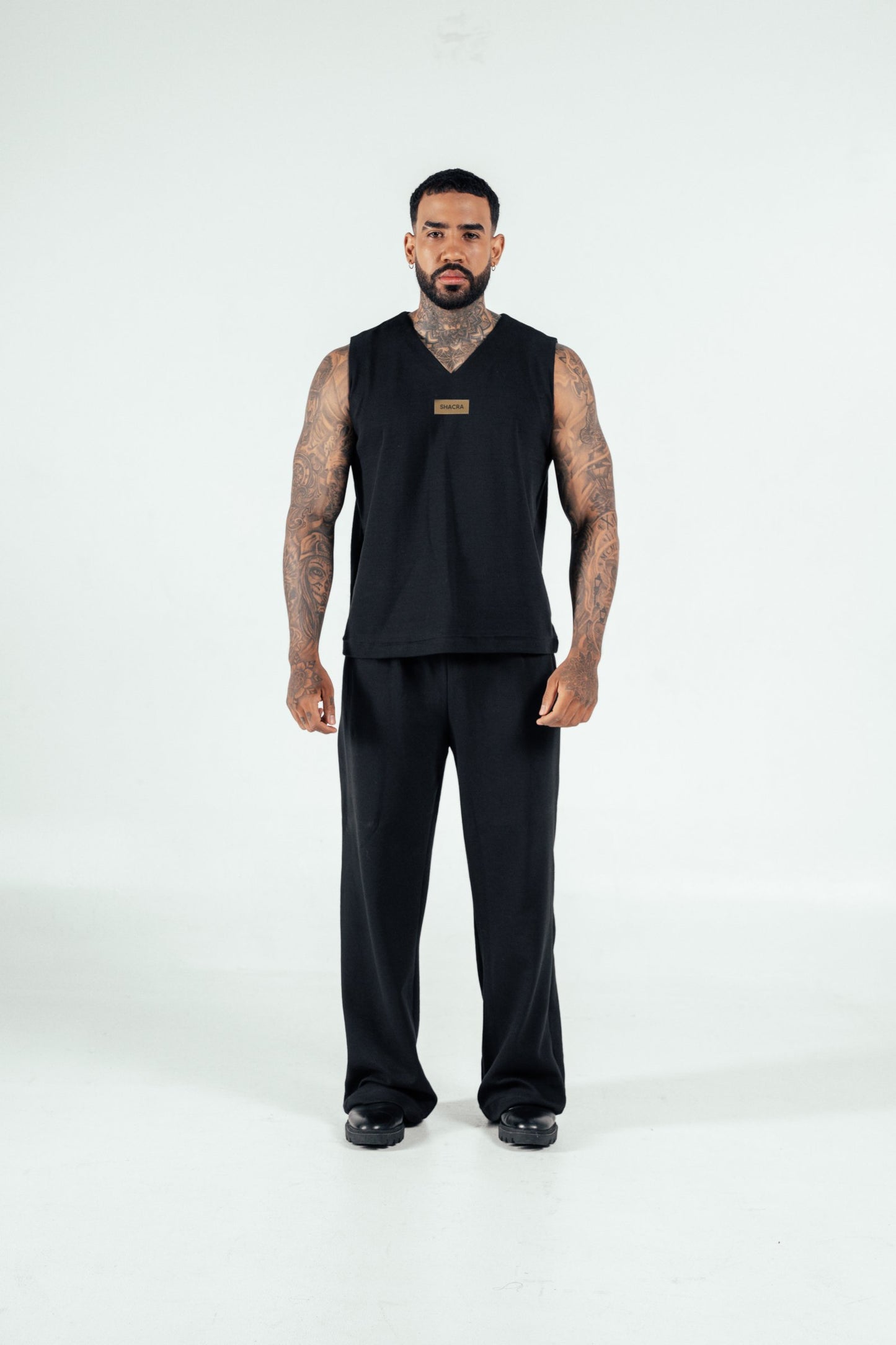Black sportswear tank top and pants with tattoos, stylish male model showcasing athletic fashion, minimalist background, modern activewear lifestyle, SHACRA branding prominently displayed.