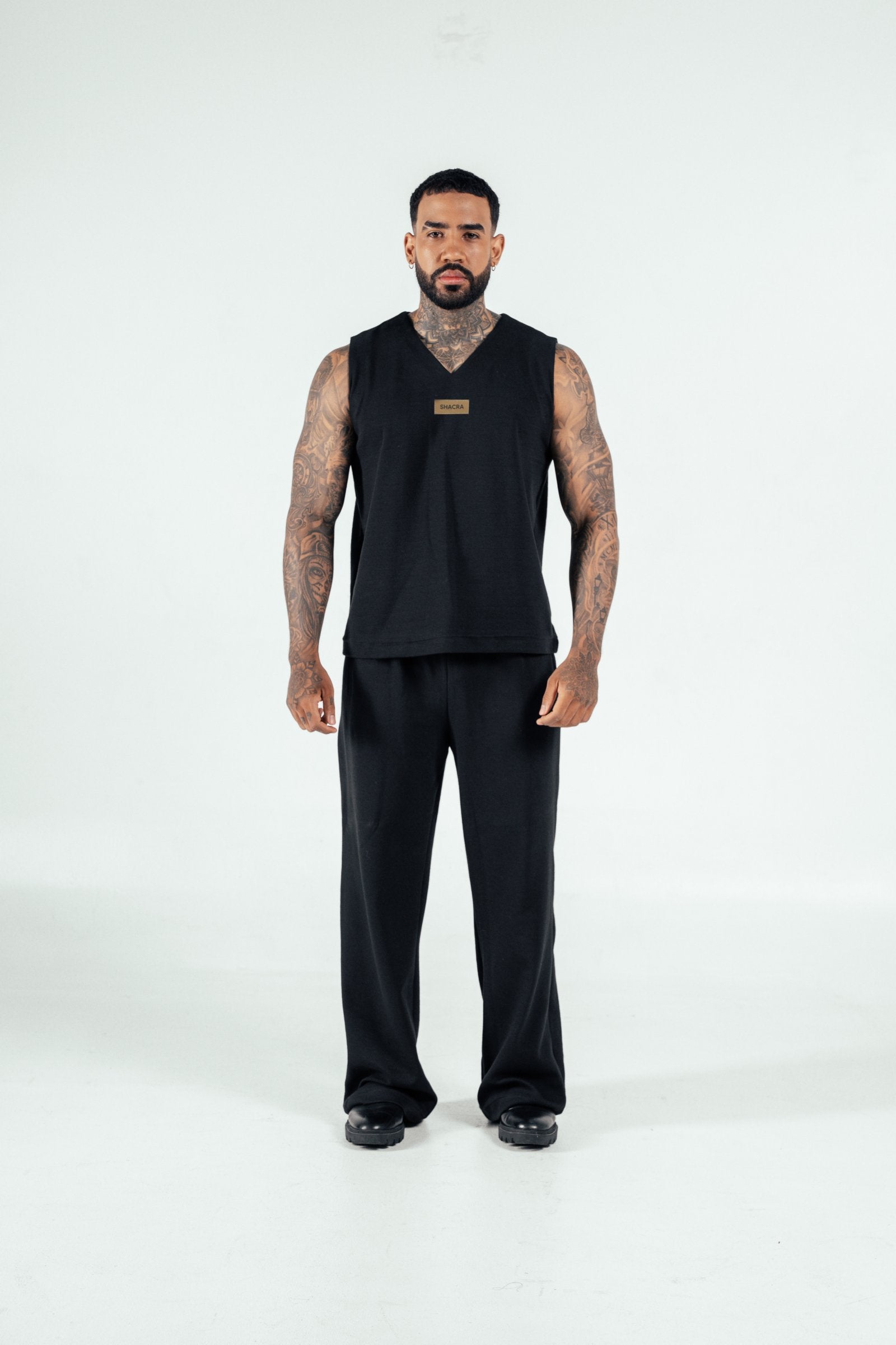 Black sportswear tank top and pants with tattoos, stylish male model showcasing athletic fashion, minimalist background, modern activewear lifestyle, SHACRA branding prominently displayed.
