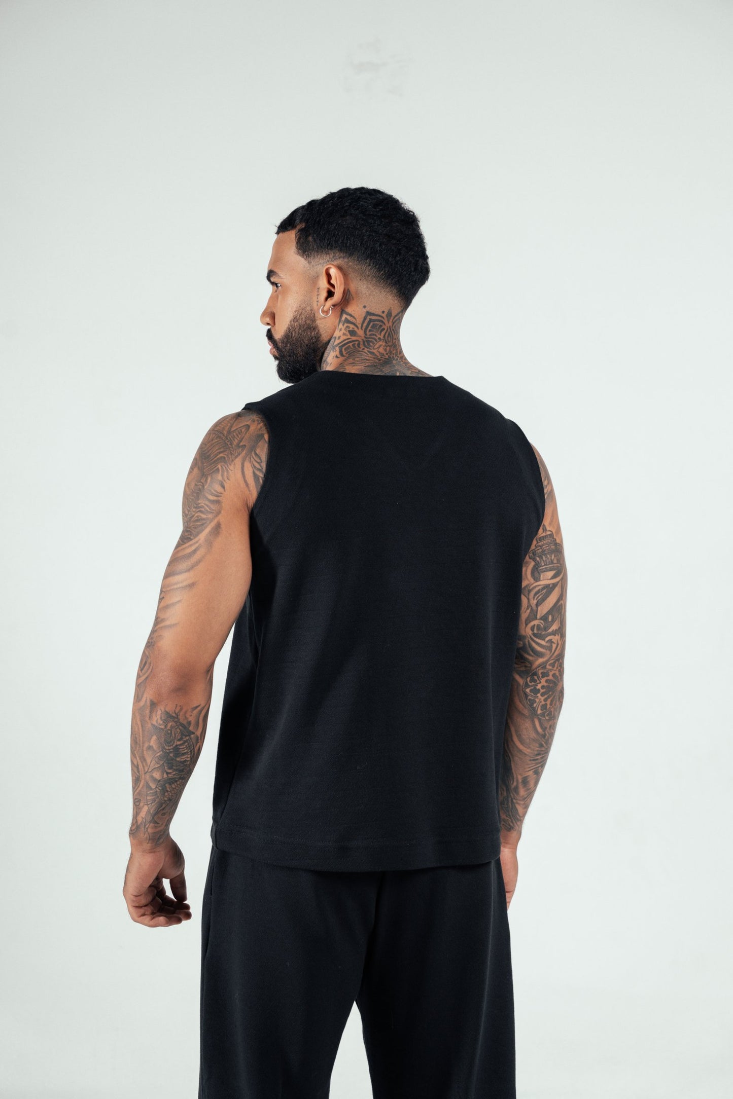 Tattooed man wearing black sleeveless top and black pants, side view, modern fashion, body art, studio photography for fashion blog.