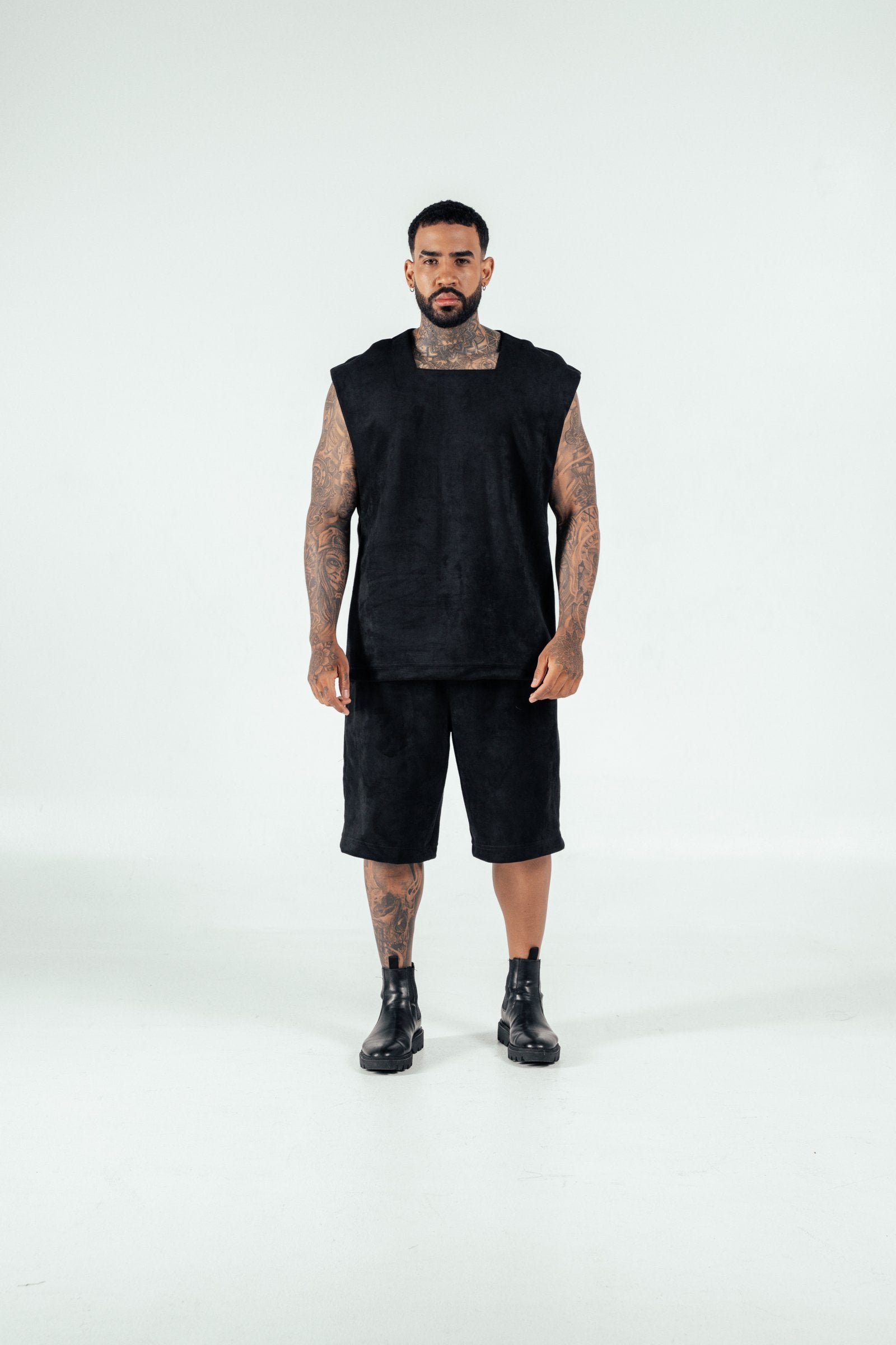 Bold tattooed man wearing black sleeveless top, shorts, and combat boots in studio against white background, showcasing edgy fashion style and body art.