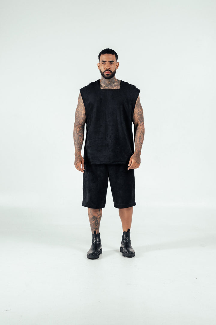 Bold tattooed man wearing black sleeveless top, shorts, and combat boots in studio against white background, showcasing edgy fashion style and body art.