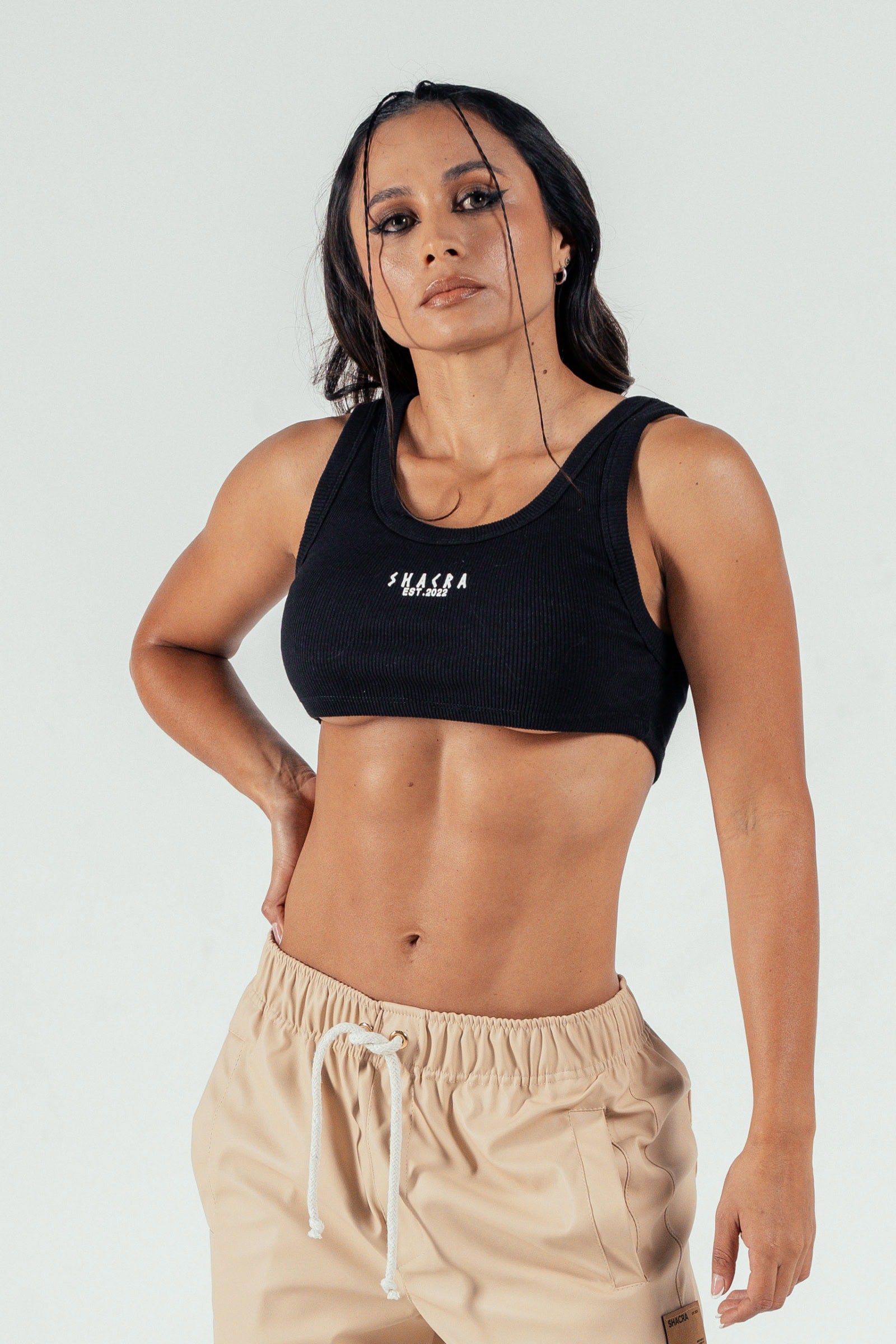 Elegant woman modeling casual athleisure wear, showcasing stylish black crop top with "Shacra" logo and beige drawstring shorts, emphasizing trendy activewear for modern lifestyle.