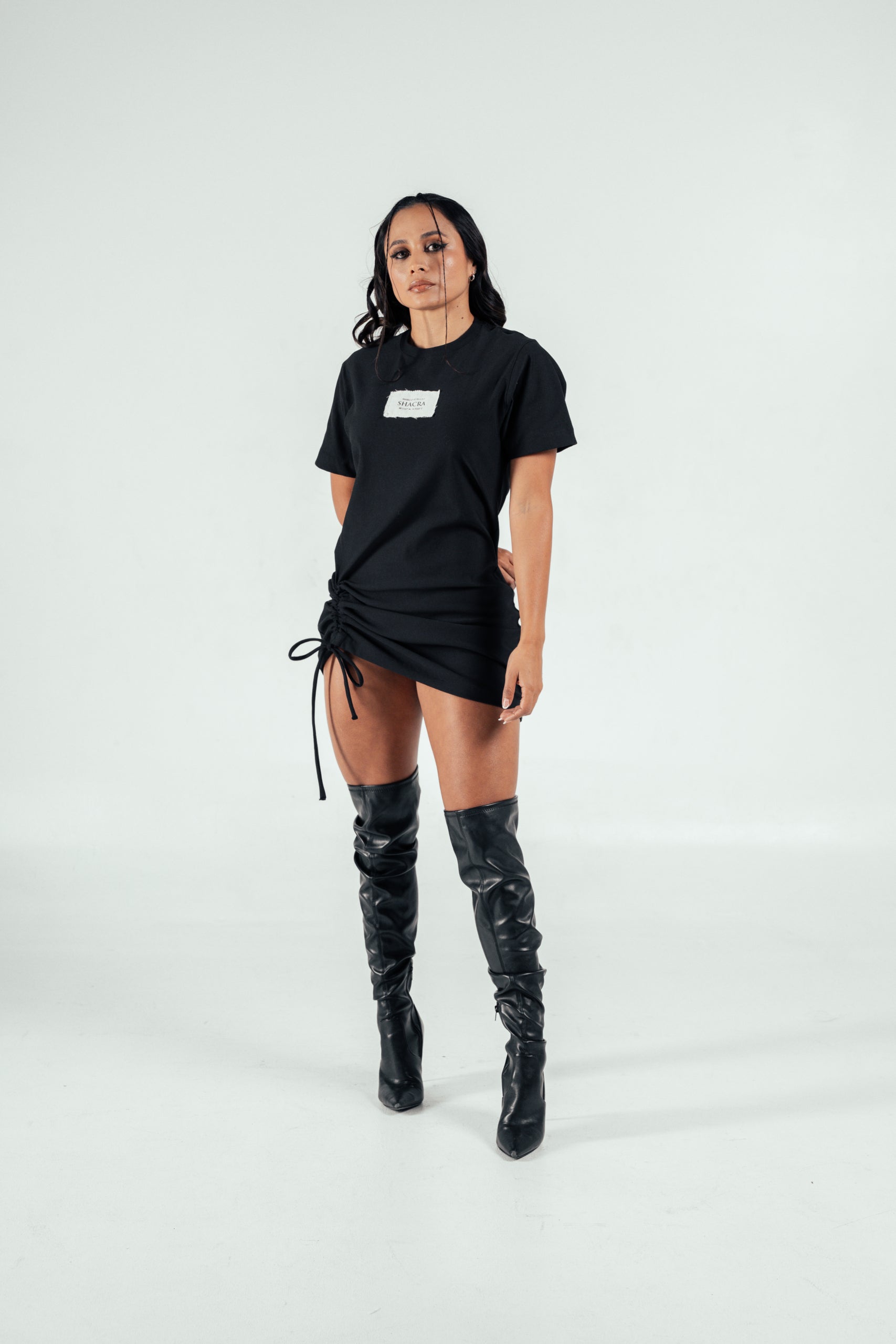 Bold woman wearing black T-shirt with "SHACRA" logo and stylish thigh-high black leather boots, posing confidently against a plain white background.