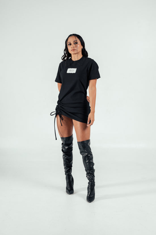 Bold woman wearing black T-shirt with "SHACRA" logo and stylish thigh-high black leather boots, posing confidently against a plain white background.