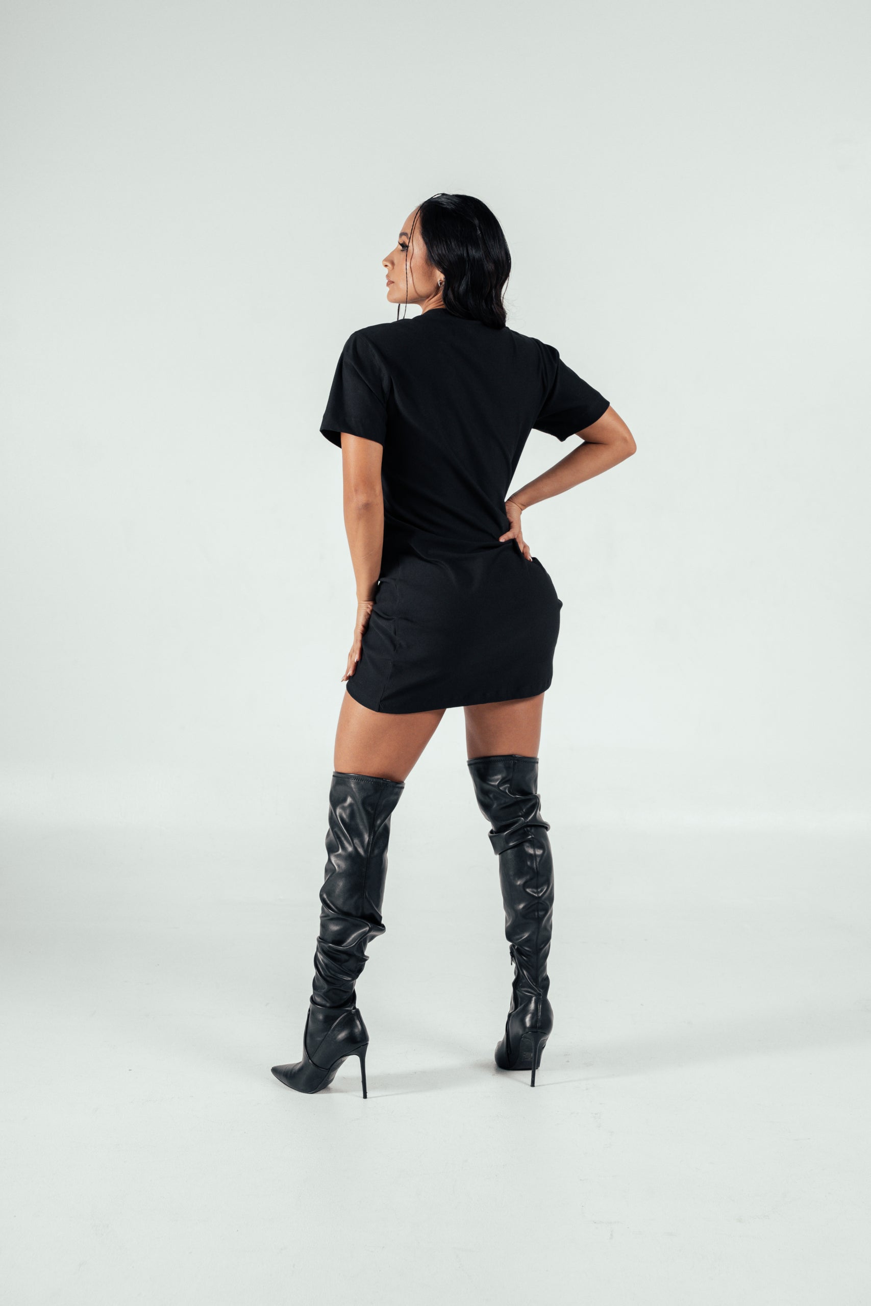 Sassy woman modeling stylish black T-shirt dress with thigh-high leather boots on a plain white background. Perfect look for fashion-forward, confident women focused on trendy urban attire.