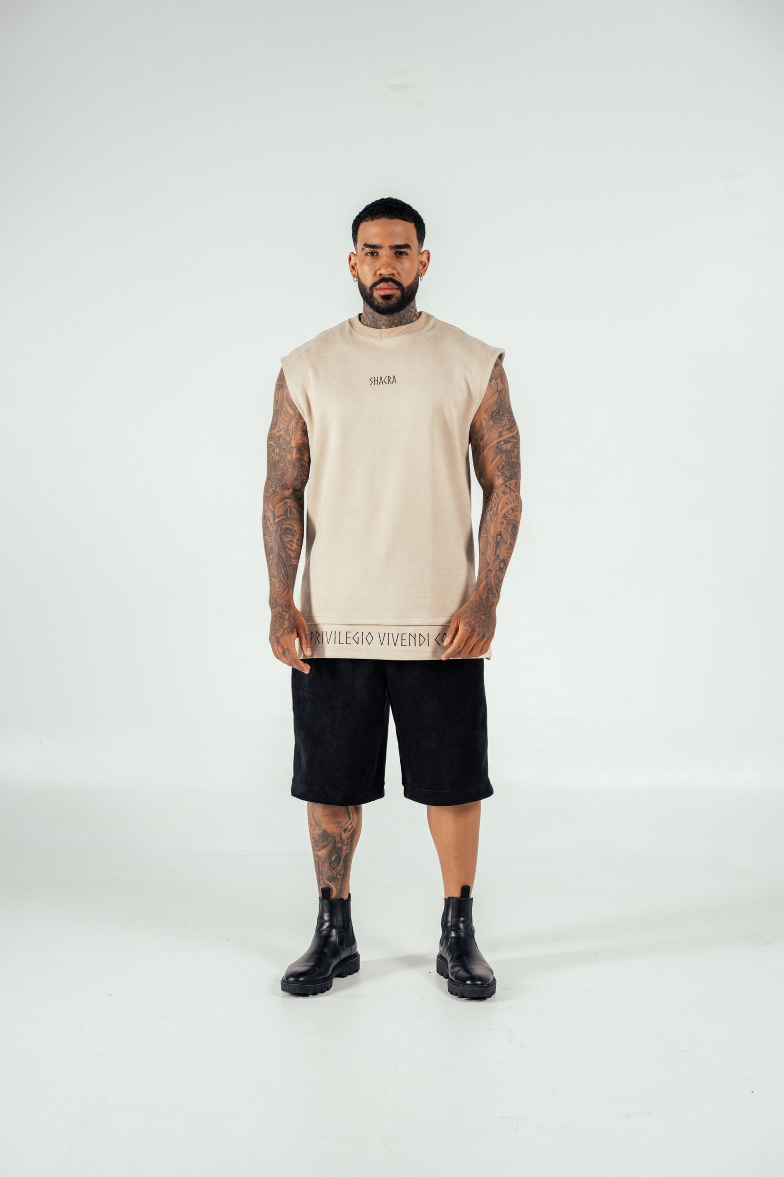 Stylish man wearing a beige sleeveless top with "Shacra" logo, black shorts, and combat boots, showcasing contemporary streetwear fashion, tattoos, and confident pose for men's clothing brand.