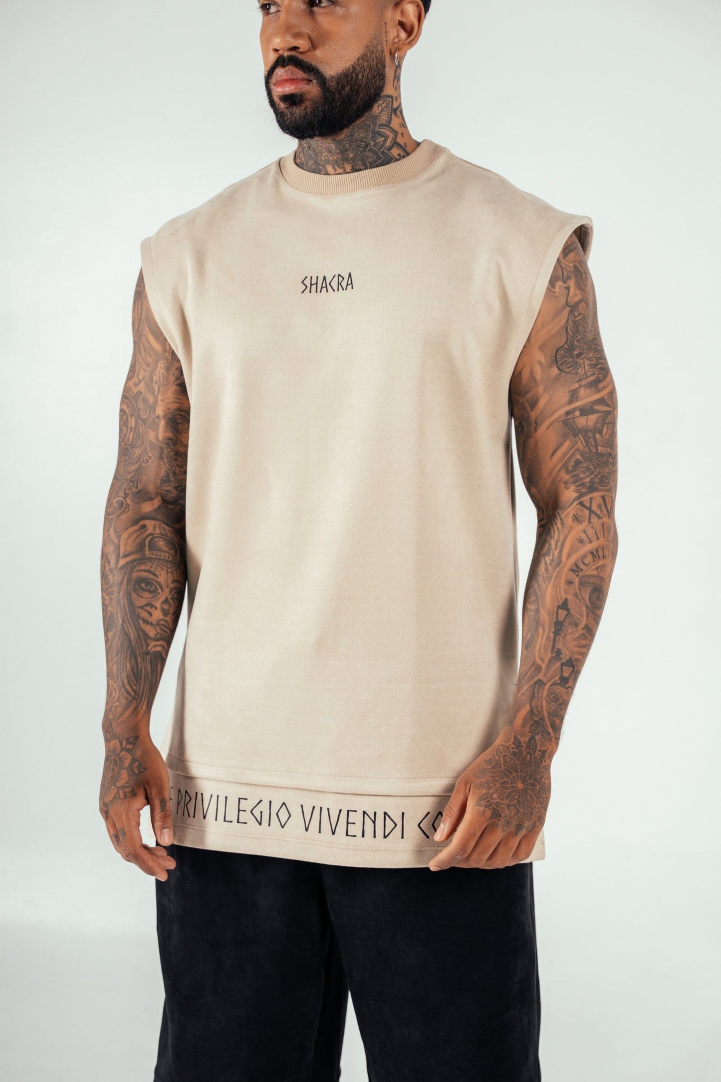 Bold tattooed male model wearing a sleeveless beige top with "SHACRA" logo, showcasing streetwear fashion, tattoos, and modern style, against a plain background.