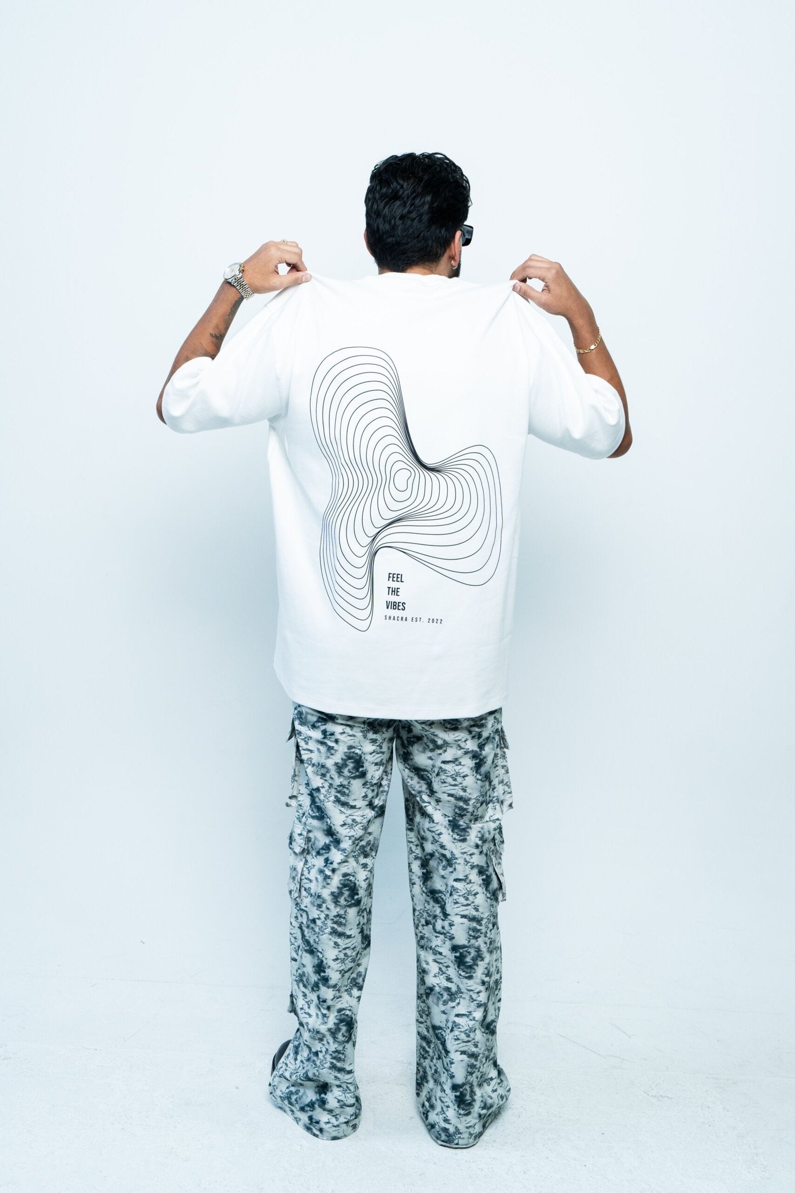 Modern man wearing graphic t-shirt from Shacra brand showcasing contemporary fashion and stylish casual wear, standing against a plain white background, highlighting the apparel design and aesthetic appeal.
