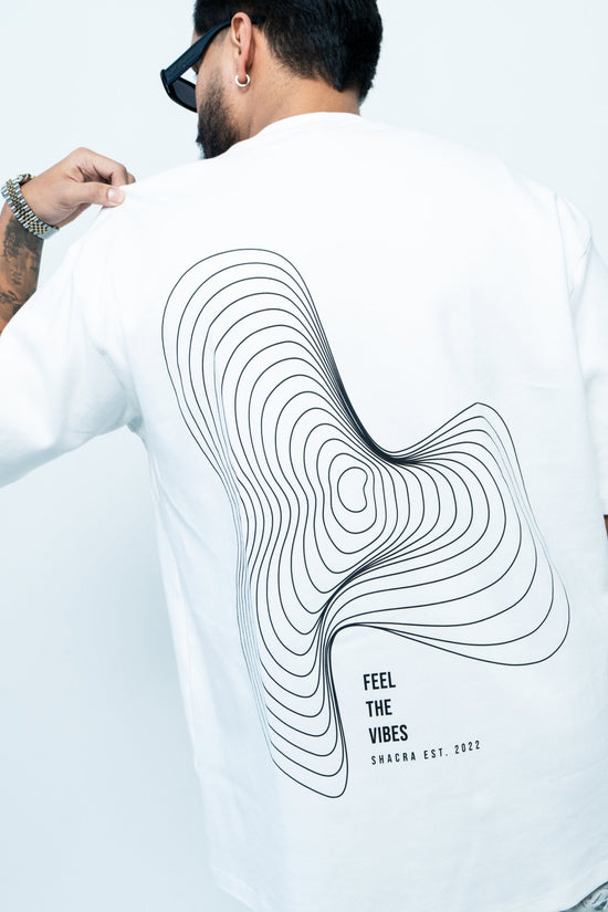 Relaxed male model wearing a white T-shirt with abstract black line art, "Feel the Vibes" slogan, and "Shacra" branding, emphasizing contemporary fashion and streetwear style.