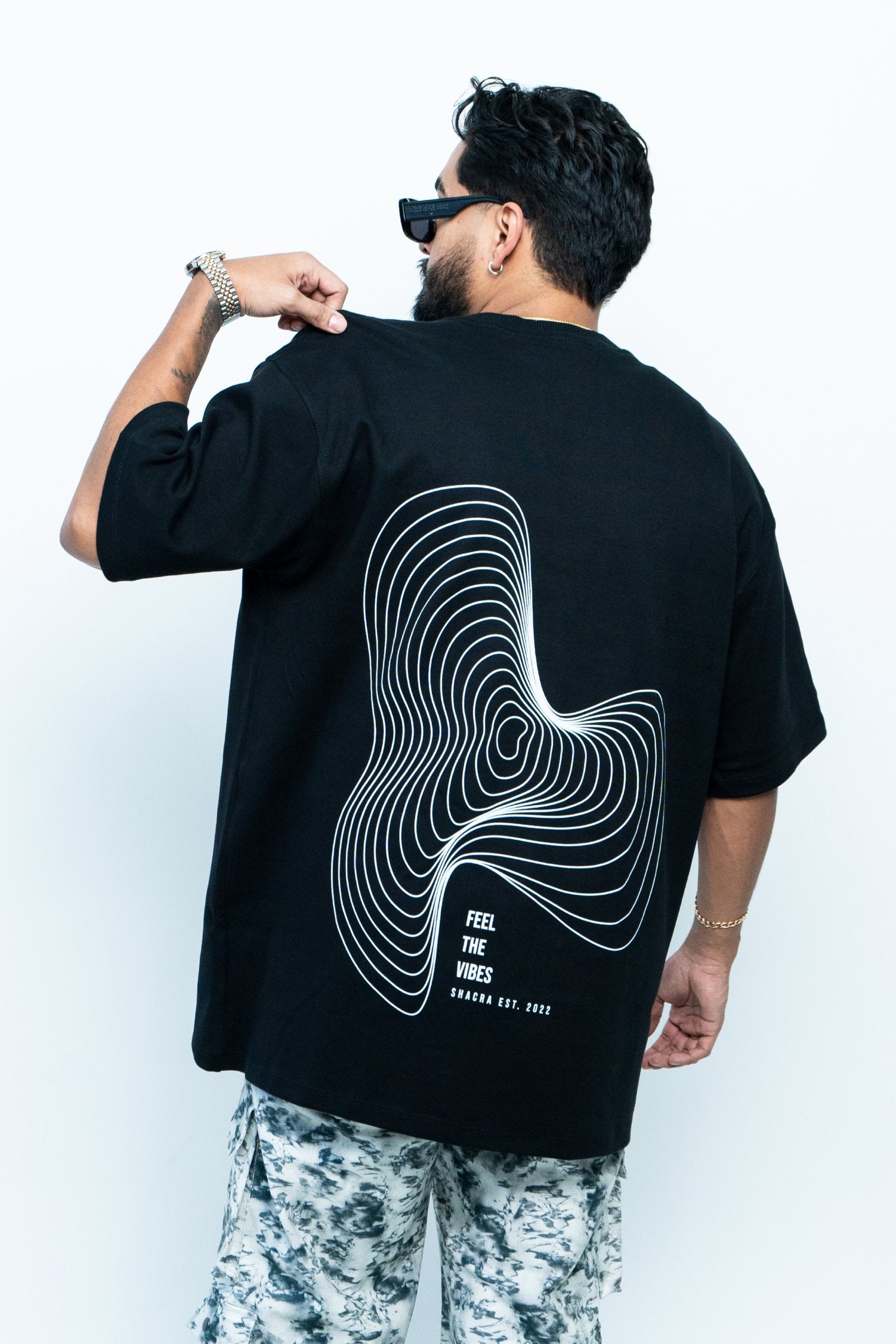 Bold black T-shirt featuring a white contour line abstract design and the phrase "Feel the Vibes" promoting Shacra, a trendy fashion brand established in 2022.