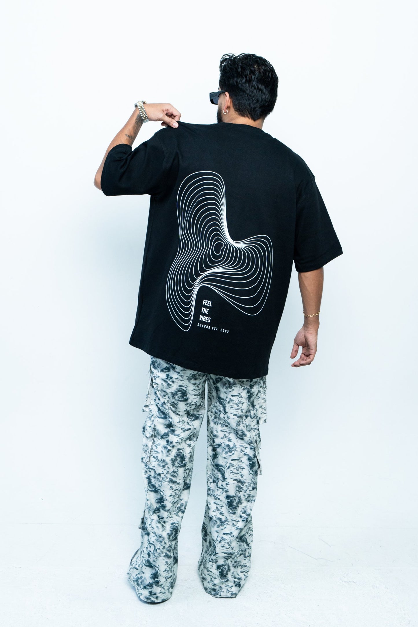Dynamic fashion photo of a person wearing a black graphic T-shirt with the text "Feel the Vibes" and contour design, paired with stylish camo pants, showcasing modern streetwear style.