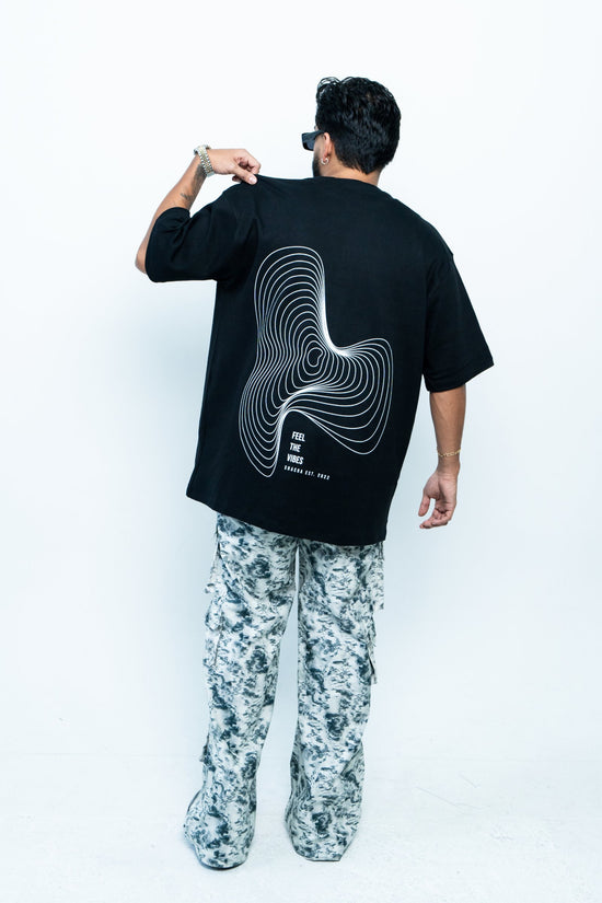 Dynamic fashion photo of a person wearing a black graphic T-shirt with the text "Feel the Vibes" and contour design, paired with stylish camo pants, showcasing modern streetwear style.