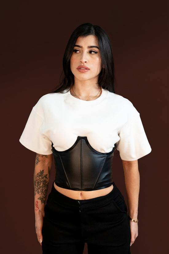 Bold woman wearing white t-shirt and black leather corset with tattooed arm against brown background.