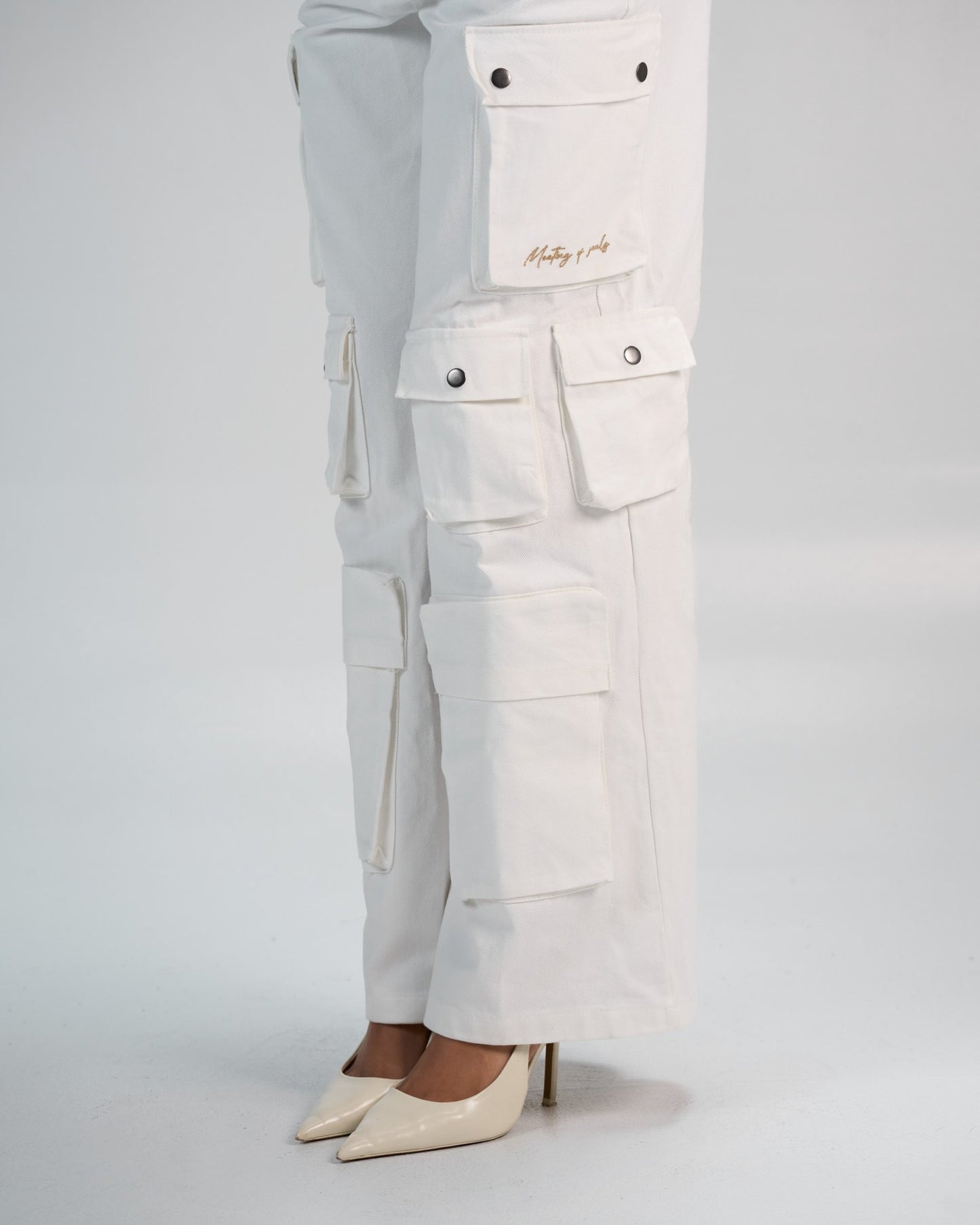 White cargo pants with multiple pockets and embroidered text, paired with beige high heels, featured in a fashion-forward image showcasing trendy streetwear style.
