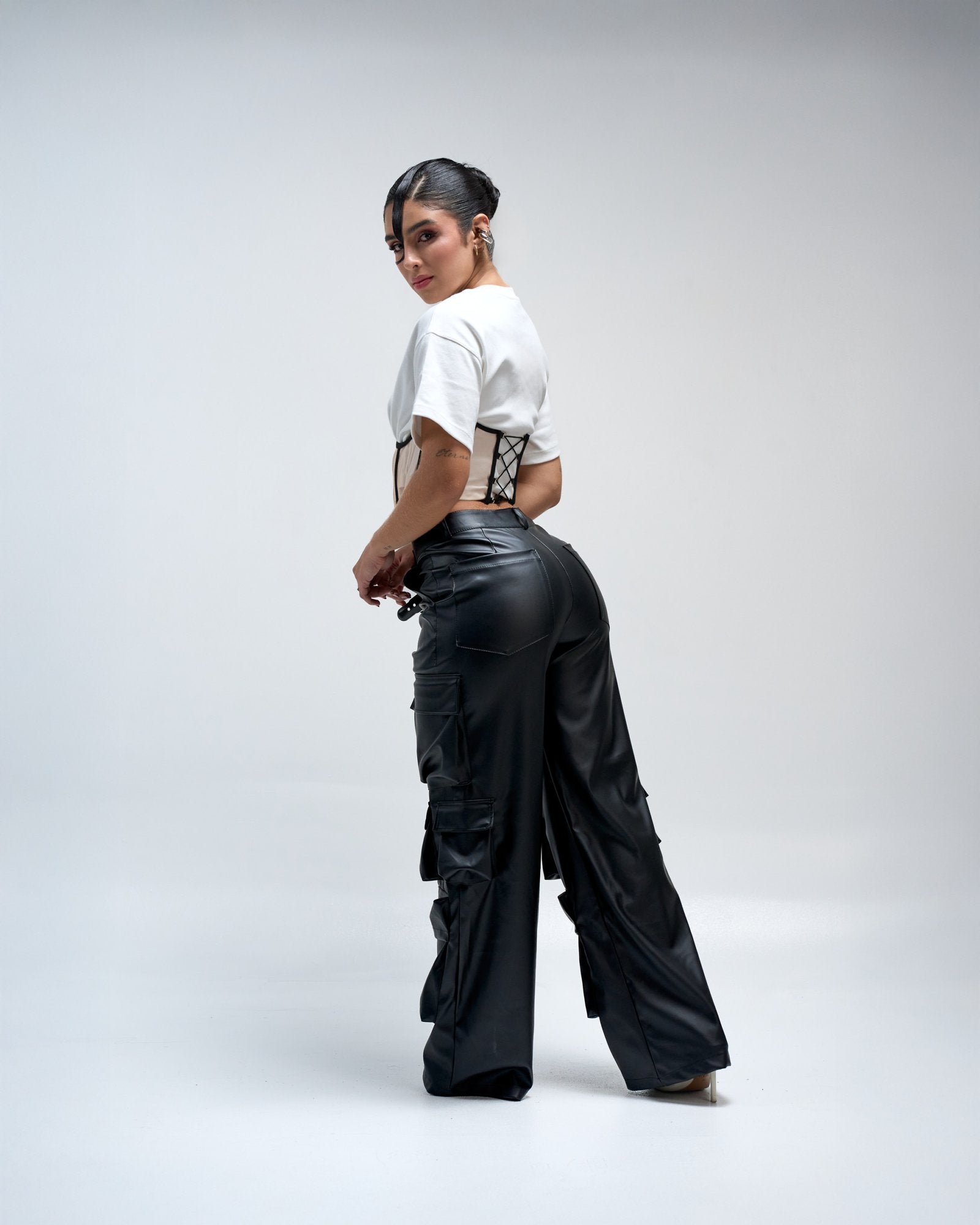 Stylish woman modeling black leather cargo pants and white cropped t-shirt against a plain light gray background, showcasing trendy fashion, urban streetwear, and modern clothing styles.