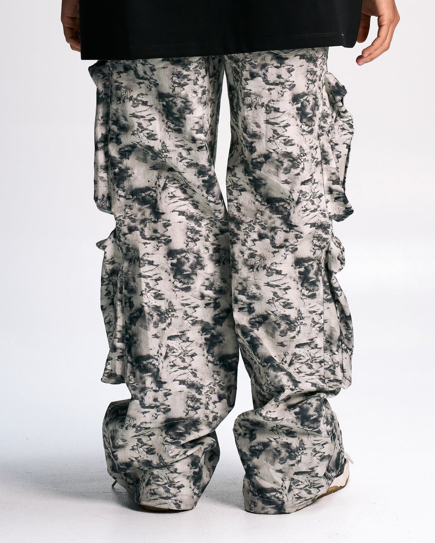 Baggy cargo pants with a black, white, and gray abstract print, featuring multiple pockets and ruffle details on the sides, styled for a fashion-forward streetwear look.